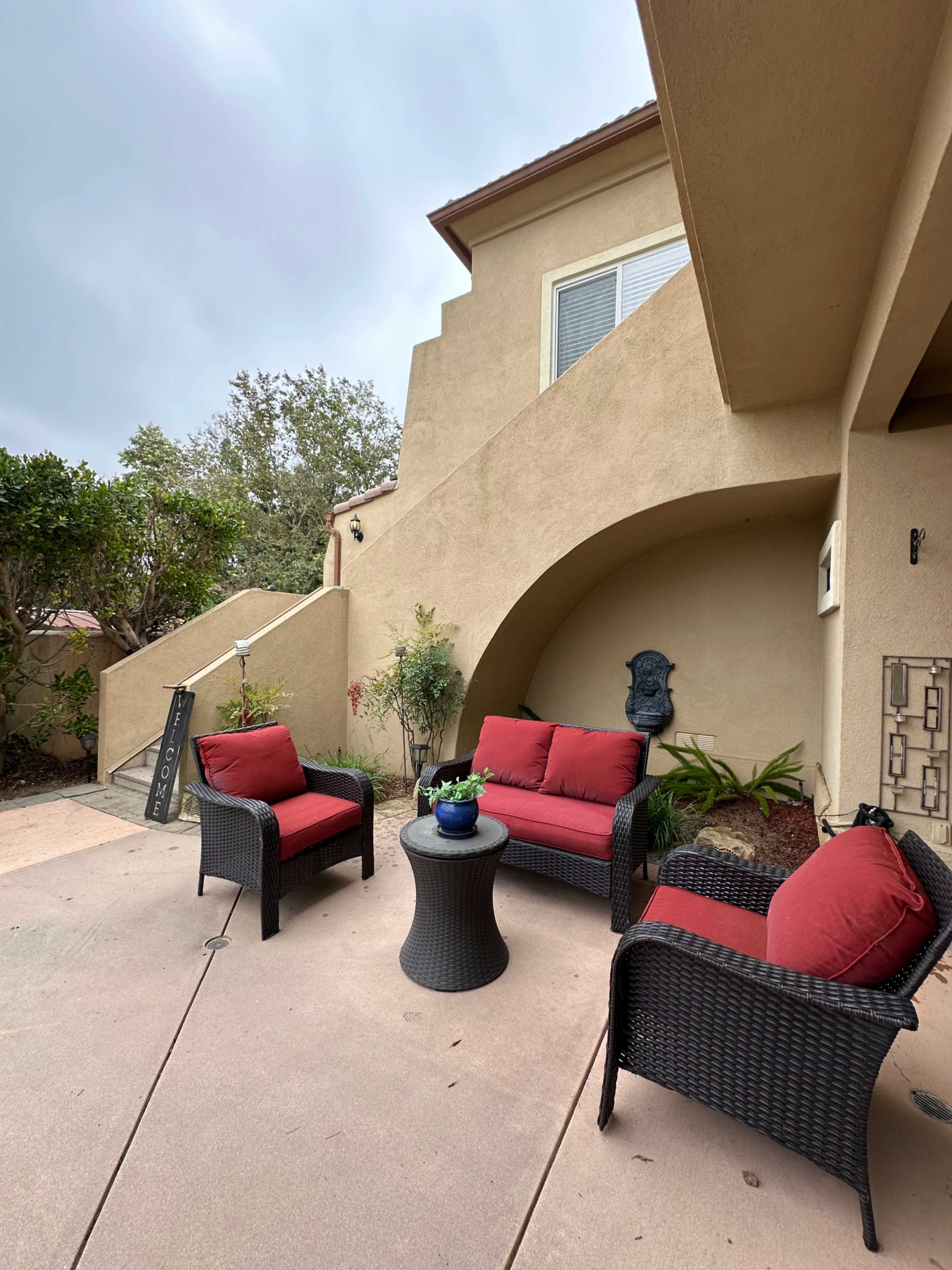 A covered patio features a set of red cushioned chairs and a small table surrounded by potted plants and a decorative wall piece near a staircase leading to an upper level.