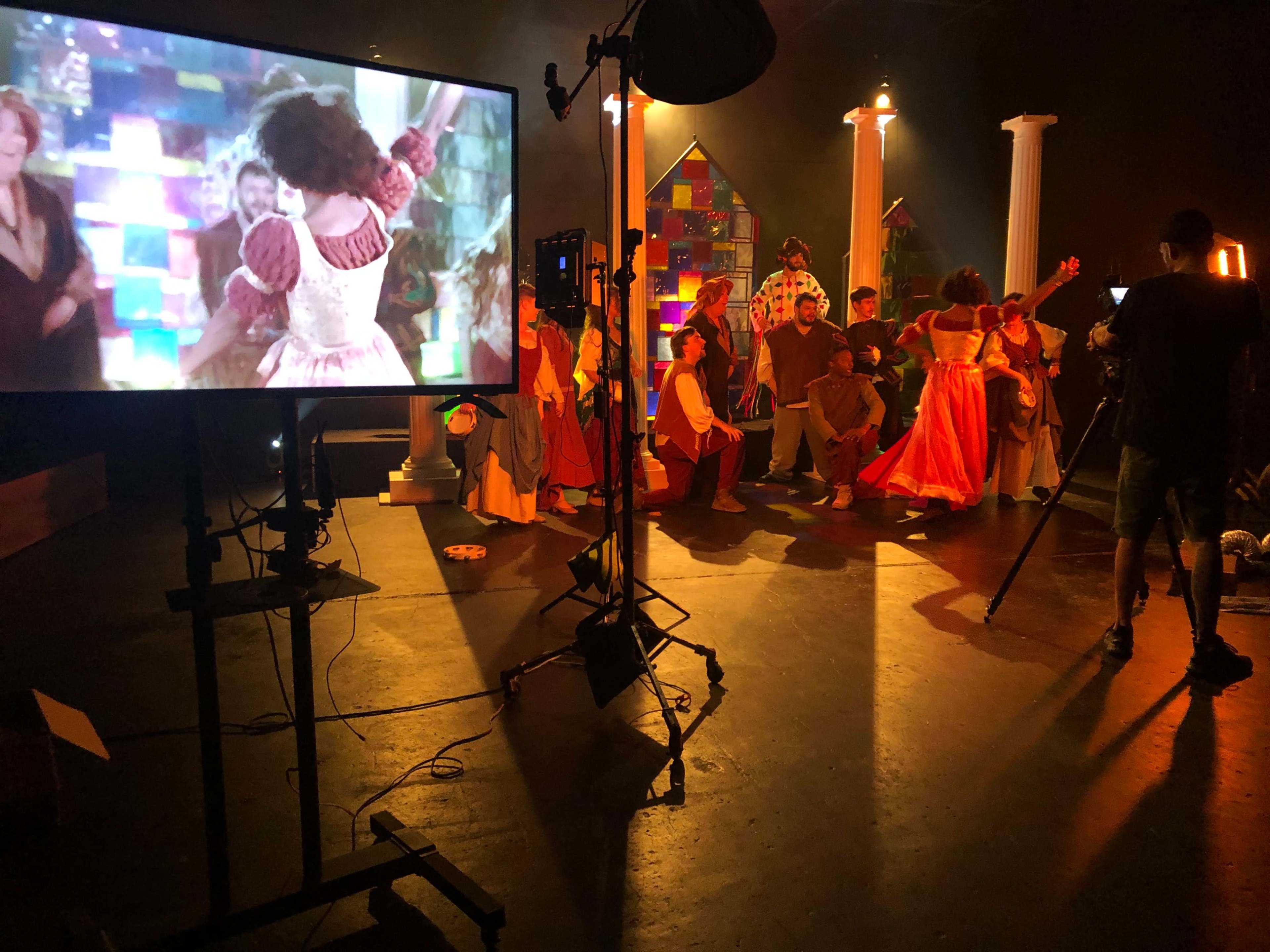 A film set features a large monitor displaying a scene, while actors in colorful costumes gather and perform in front of it.