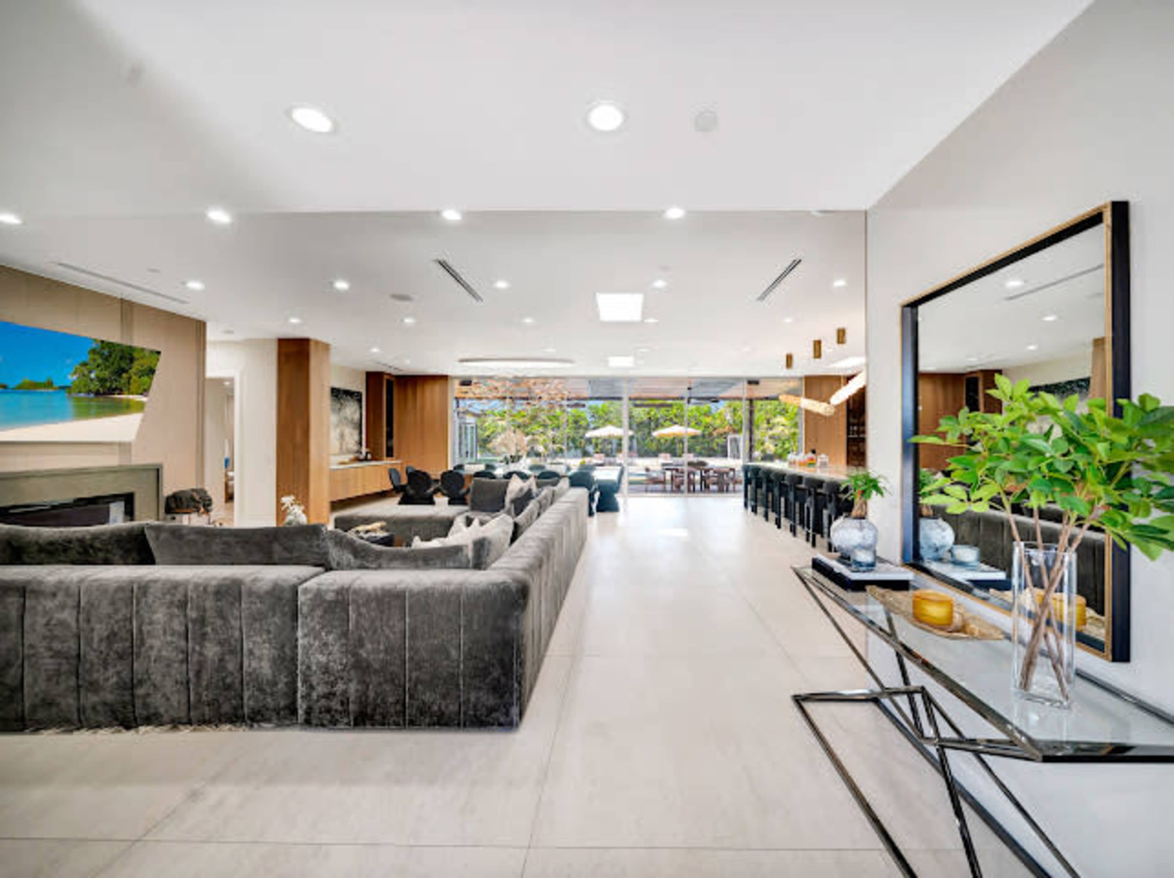 The image shows a modern living space featuring a large, gray sectional sofa, a sleek mirrored wall, and a view of an outdoor seating area through expansive glass doors.