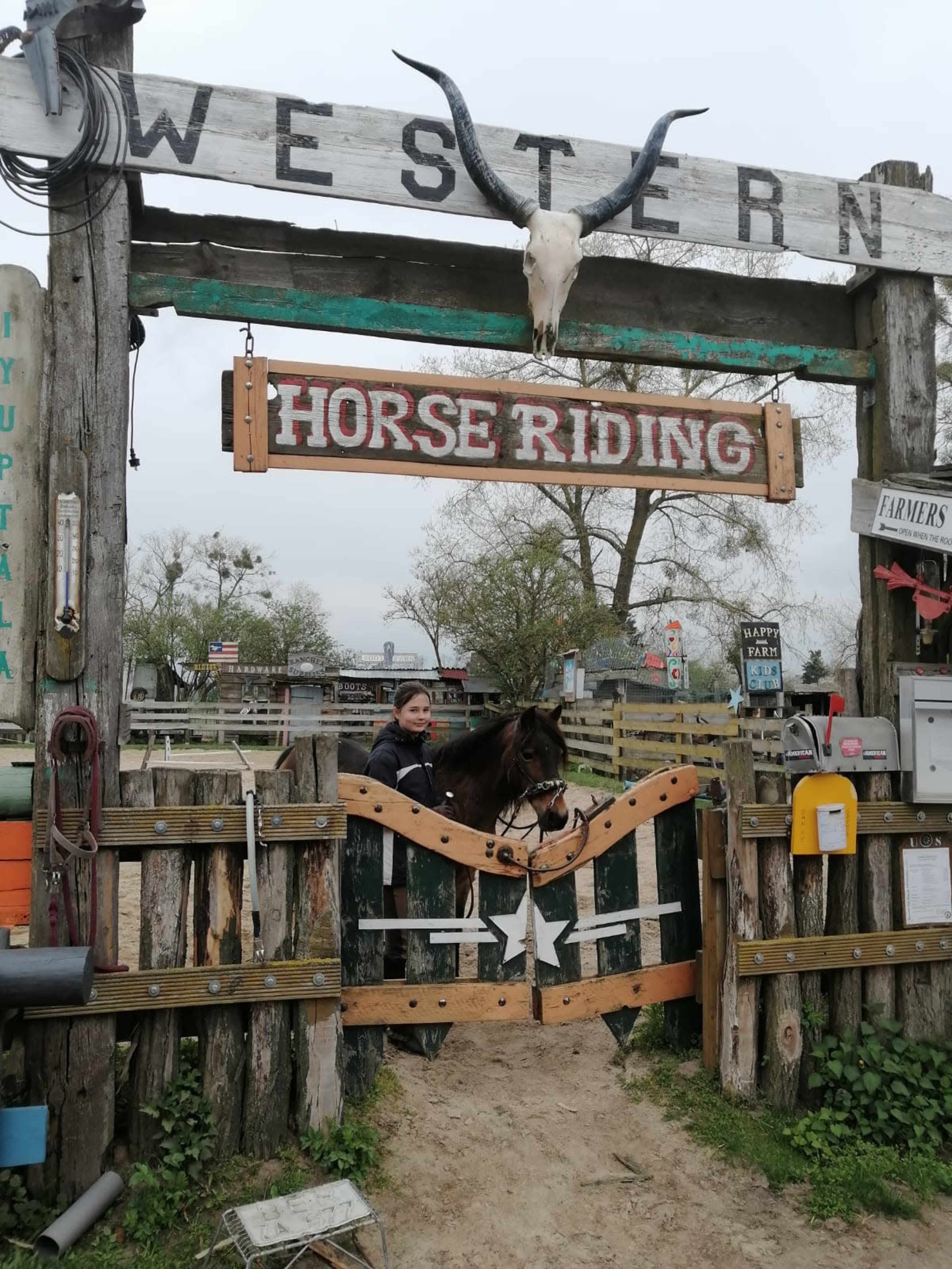 A woman leads a brown horse through a wooden gate marked "WESTERN HORSE RIDING."