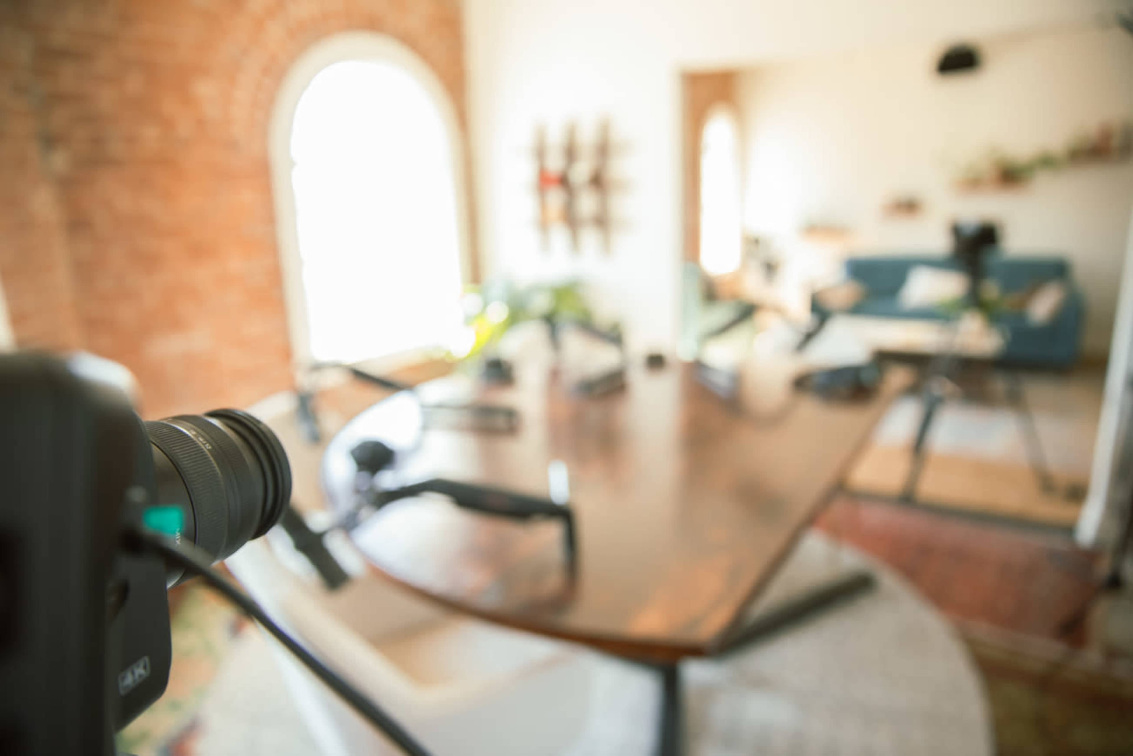 The image shows a well-lit room with a wooden table in the foreground, a camera on a tripod, and a cozy seating area in the background.