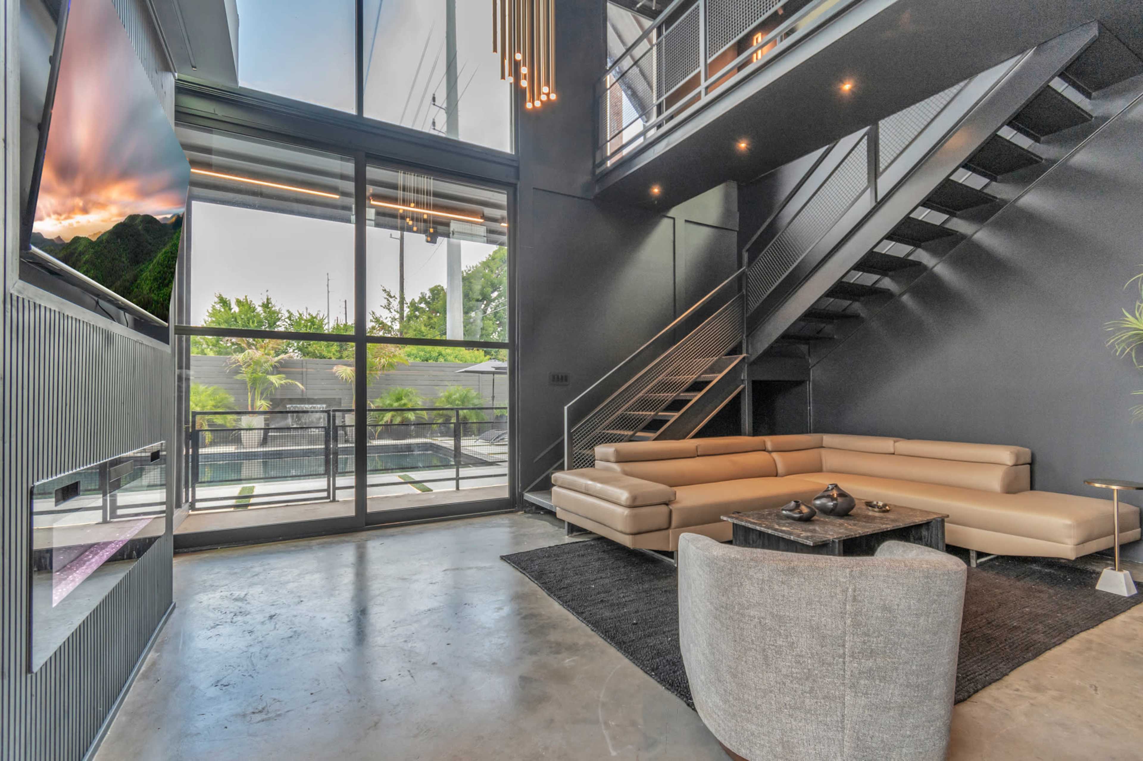 The image depicts a modern living area featuring a light-colored sectional sofa, a glass wall providing outdoor views, and a staircase leading to an upper level.