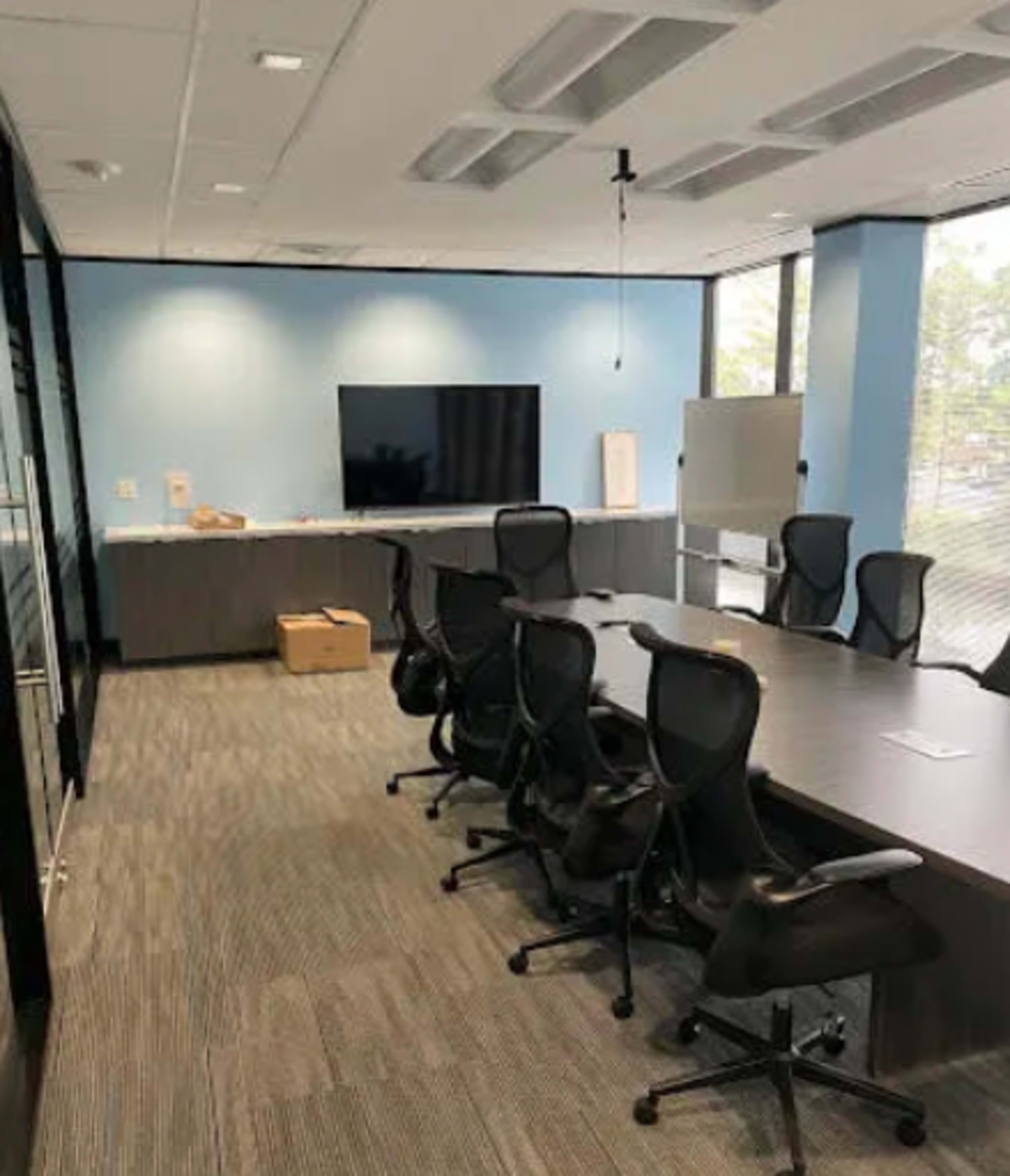 A modern conference room features a large table surrounded by ergonomic chairs, a wall-mounted television, and a whiteboard, with blue walls and windows providing natural light.