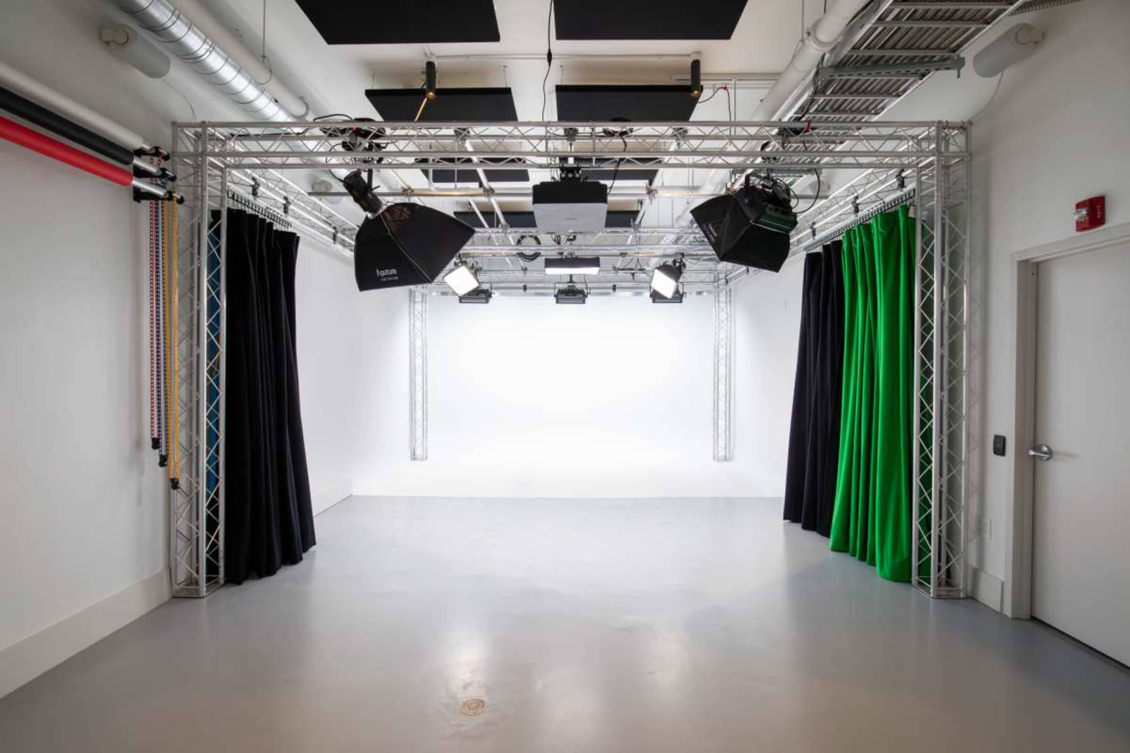 The image shows a studio space with a metal truss structure holding lights, surrounded by green and black curtains, and a white backdrop.