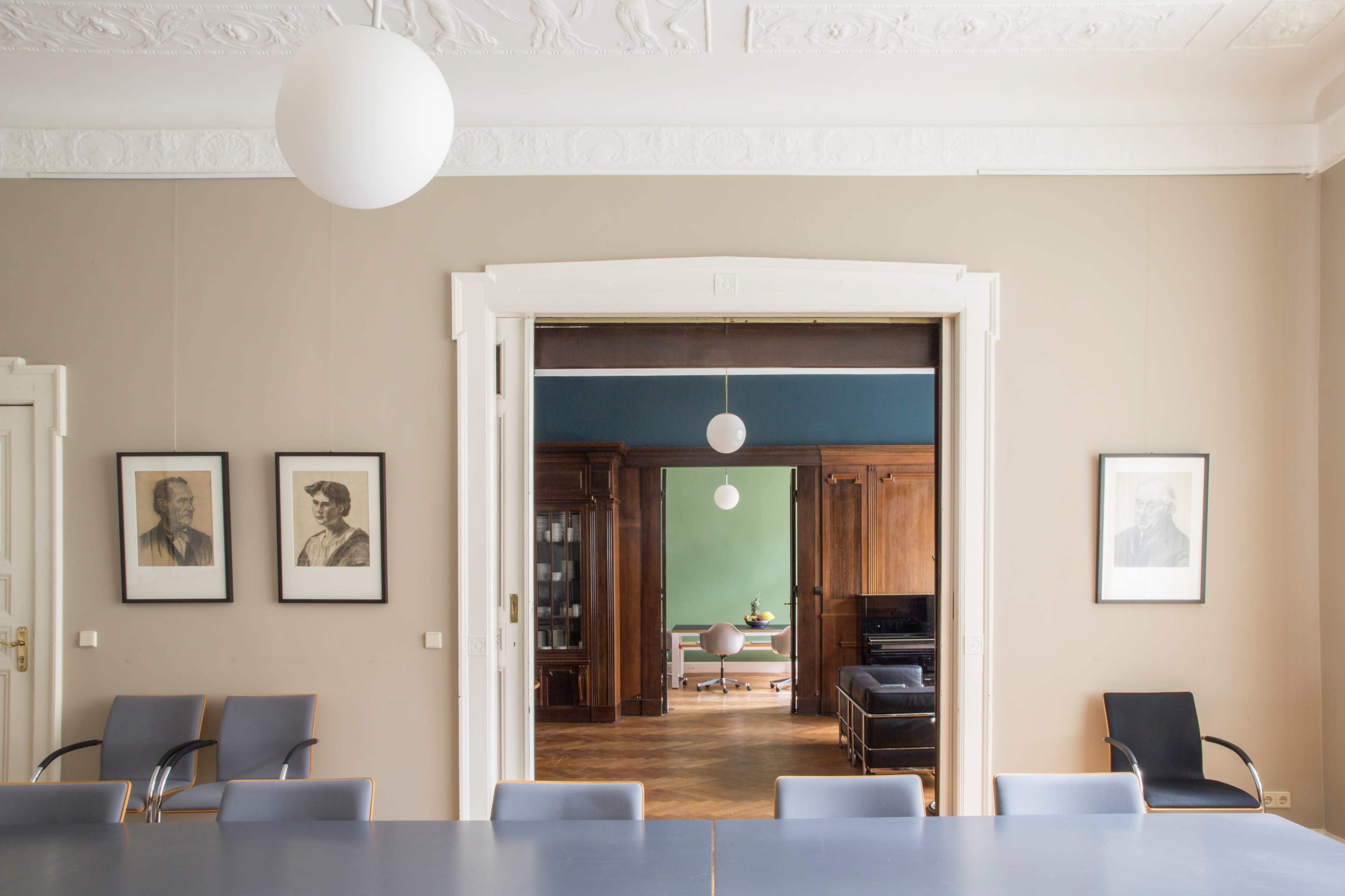 A spacious meeting room features a long table with chairs and framed portraits on the walls, leading to a brightly colored adjacent room.
