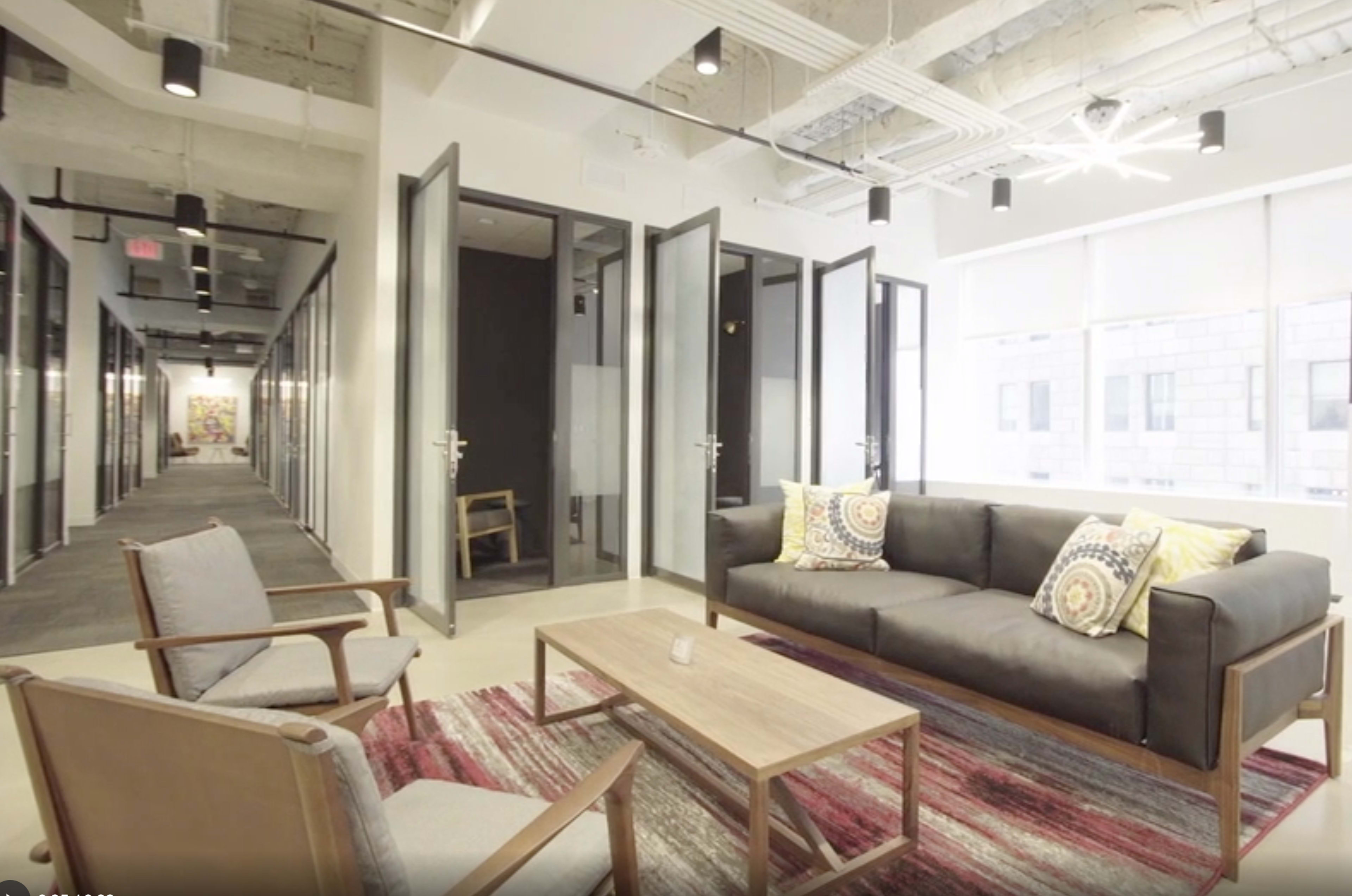 A modern office space with a lounge area featuring a sofa, armchairs, and a wooden coffee table, adjacent to glass-walled meeting rooms along a corridor.
