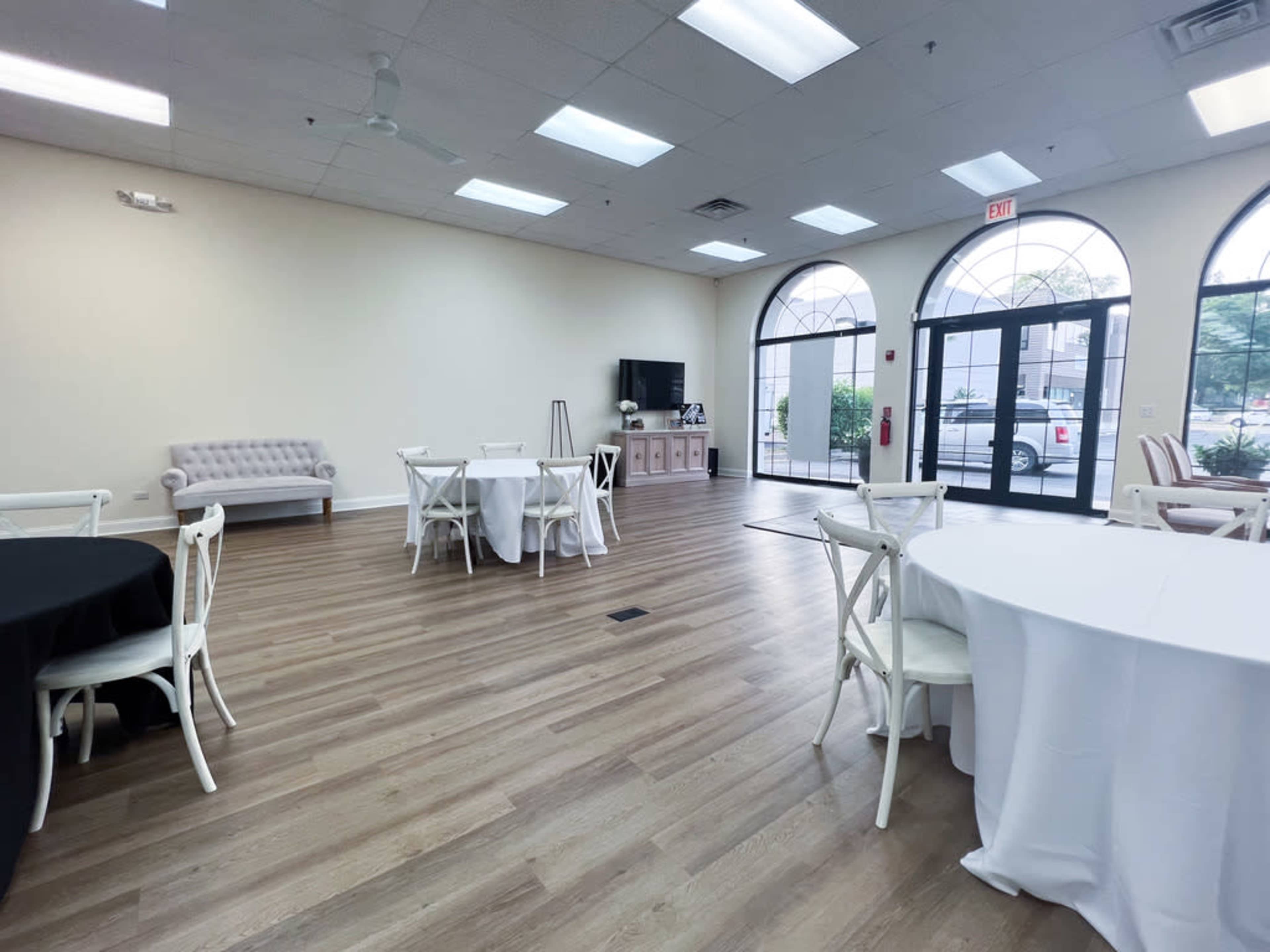 A spacious, empty room with wooden flooring, several round tables covered with white and black tablecloths, and large windows that provide natural light.