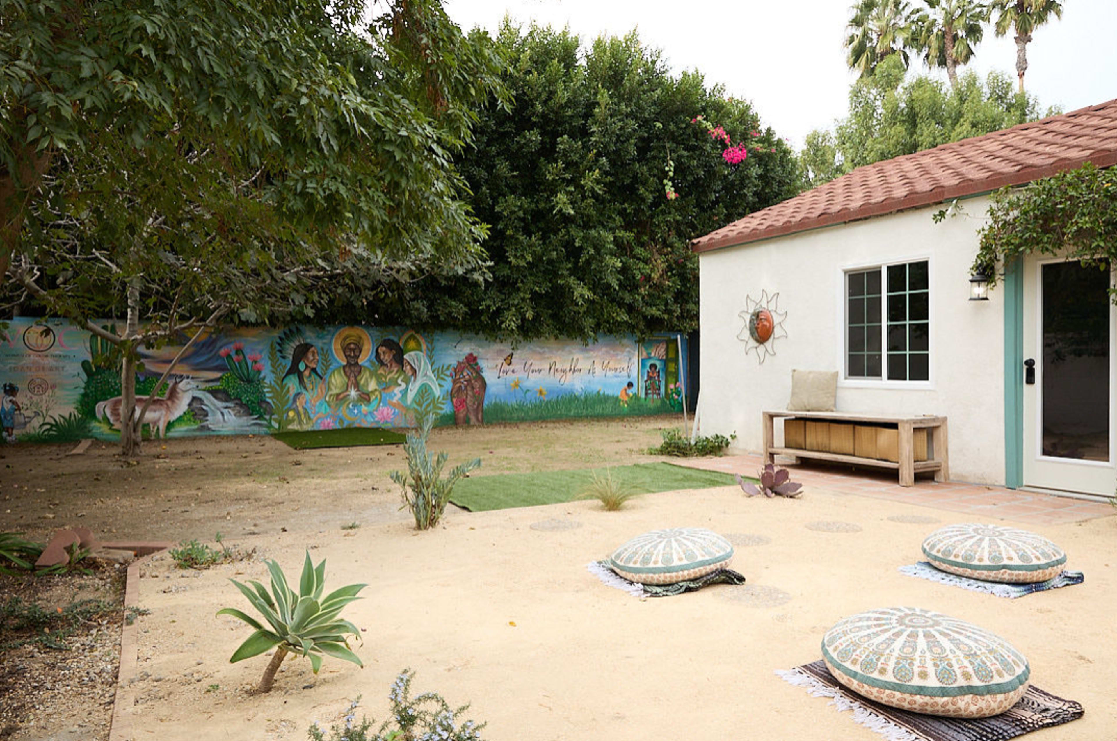 The image shows a landscaped outdoor area featuring a mural on a wall, a seating bench, and decorative poufs arranged on sandy soil.