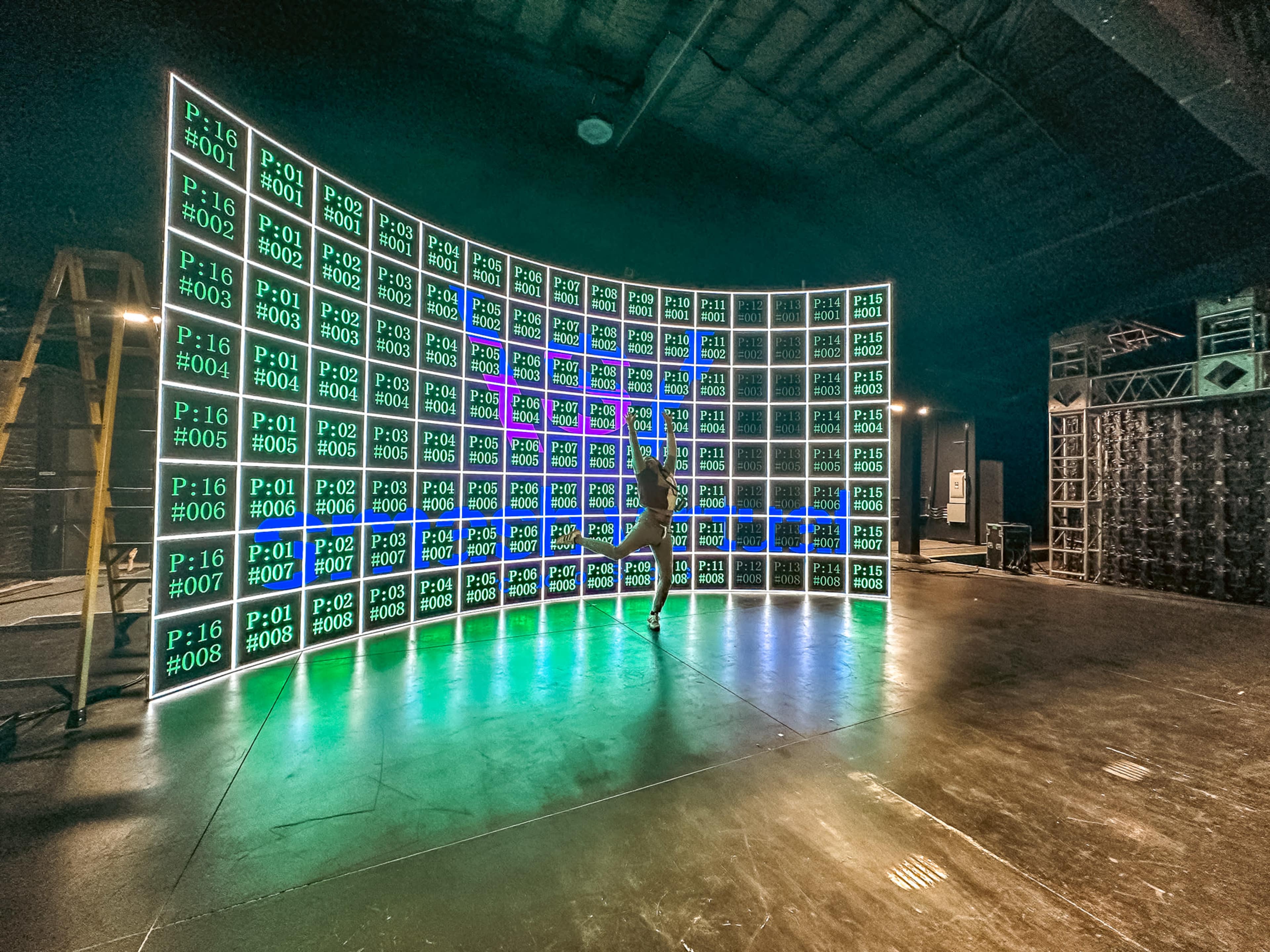 A person poses in front of a curved digital display consisting of multiple bright panels arranged neatly in a dark studio space.