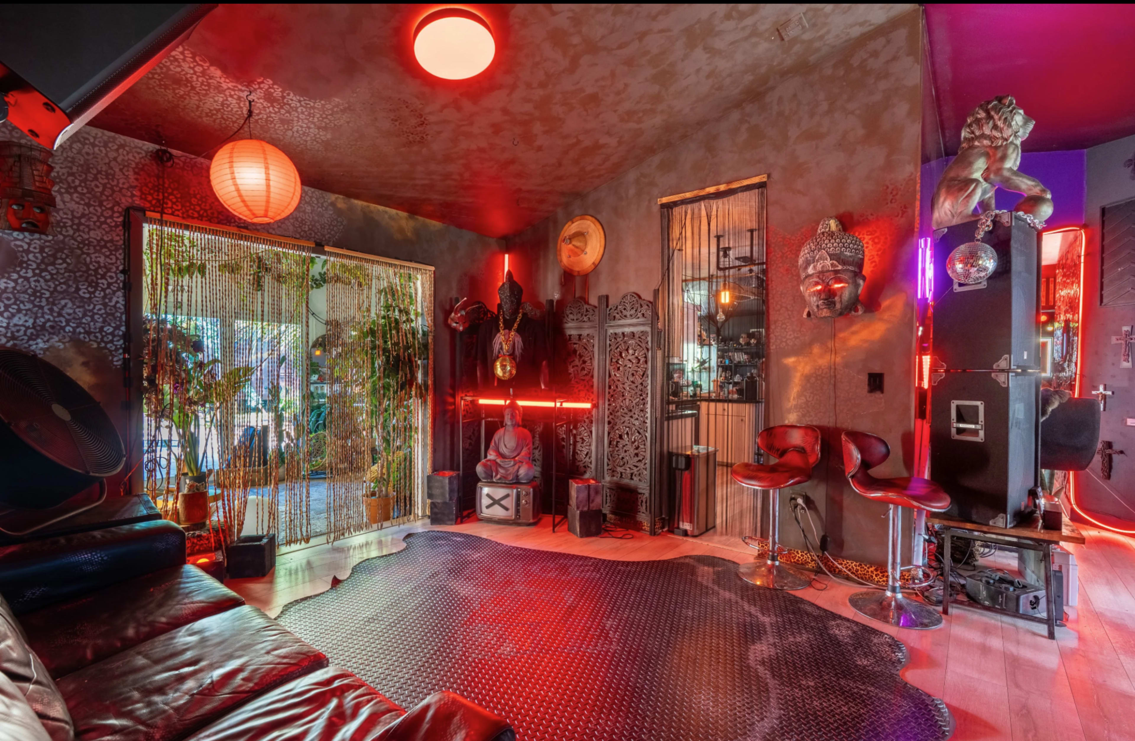 The room features eclectic decor with red lighting, a leather couch, and various statues and plants against textured walls.
