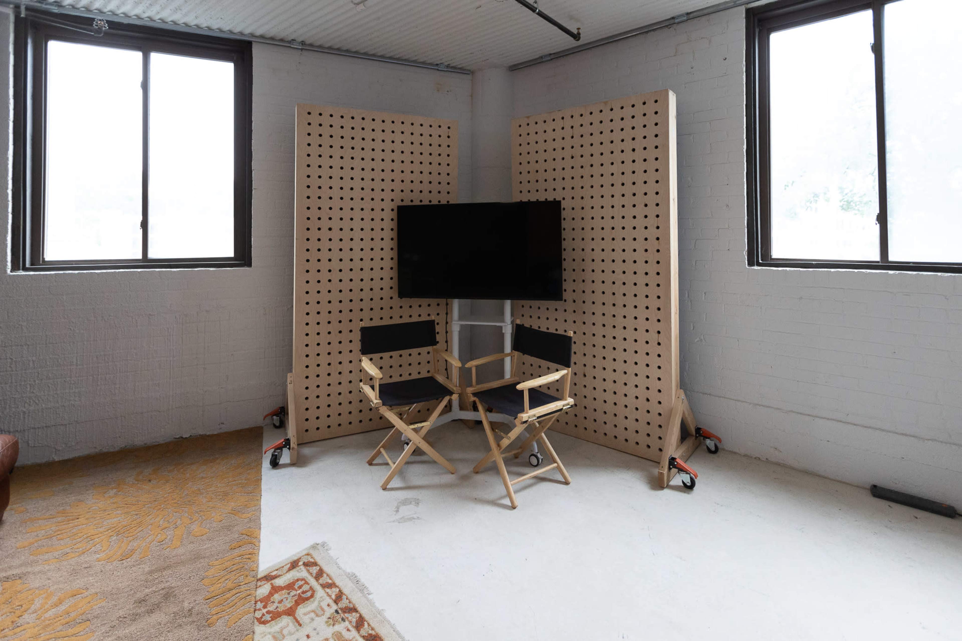 A small indoor setting with two director's chairs placed in front of a wall-mounted TV, framed by two perforated wooden partitions, with large windows on either side.