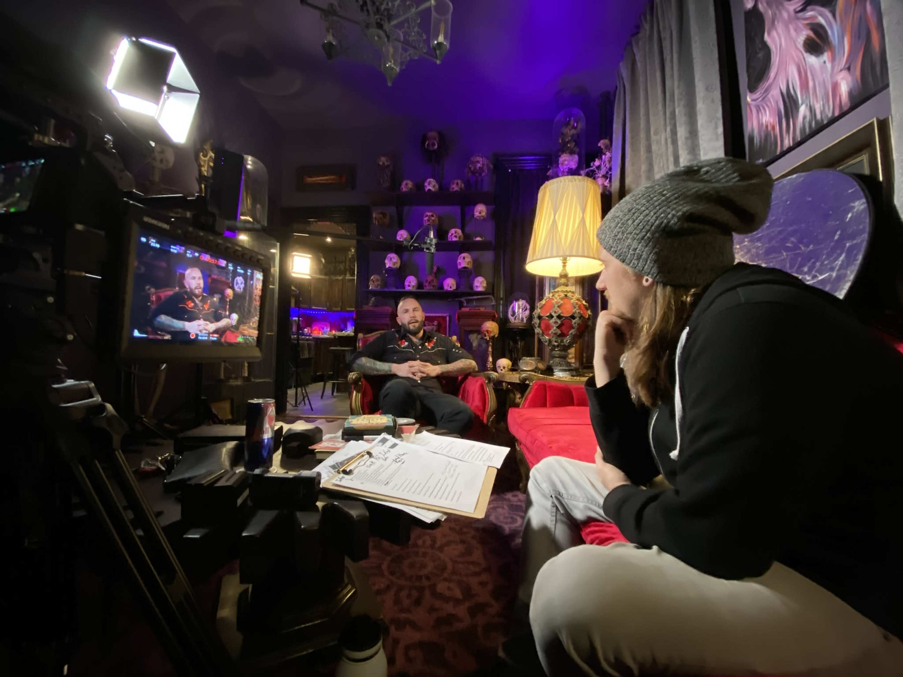 A person in a beanie sits thoughtfully while facing a well-lit interviewee on a set decorated with various curiosities and a large mirror.