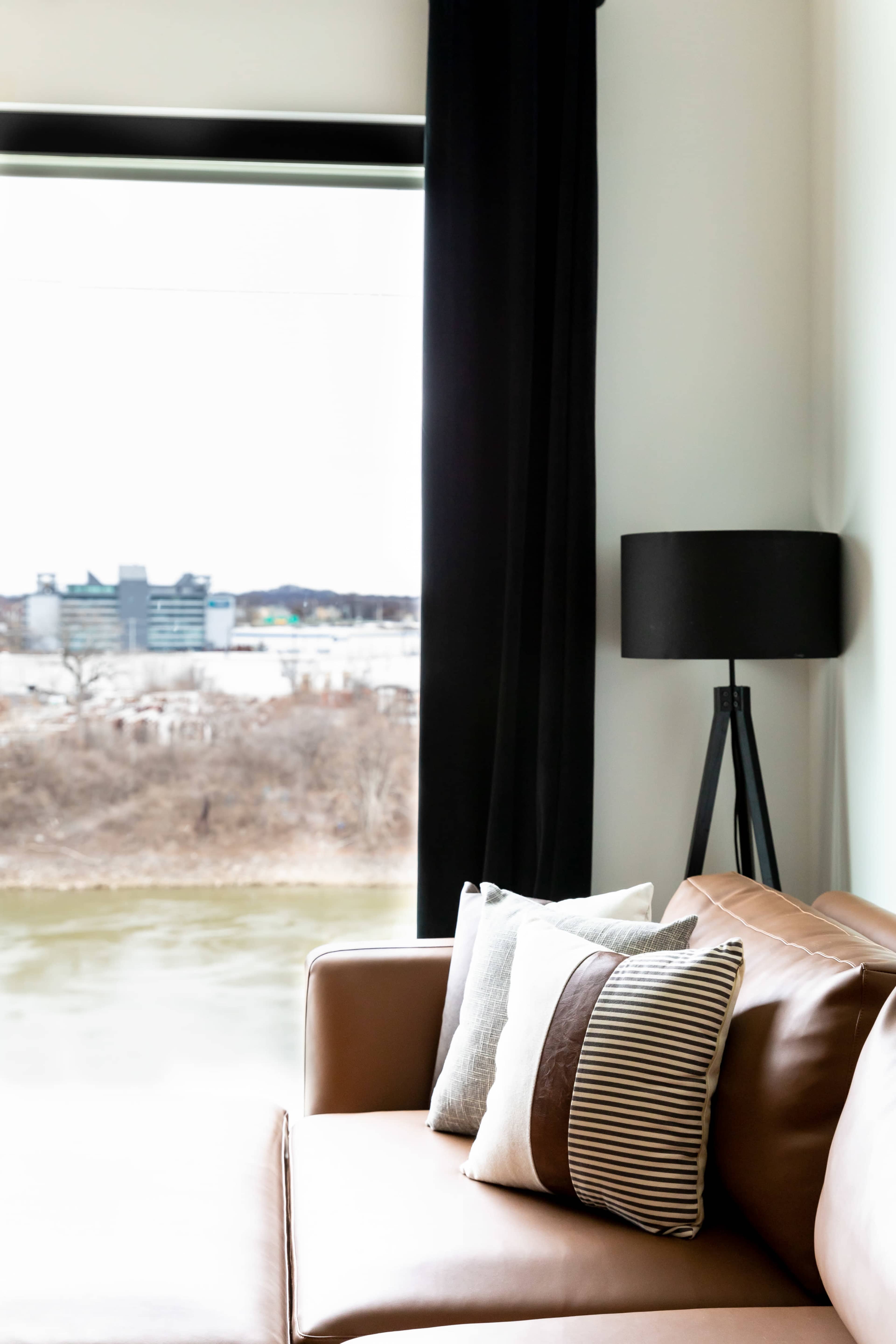 The image shows a brown couch with decorative pillows next to a floor lamp, overlooking a view through a large window.