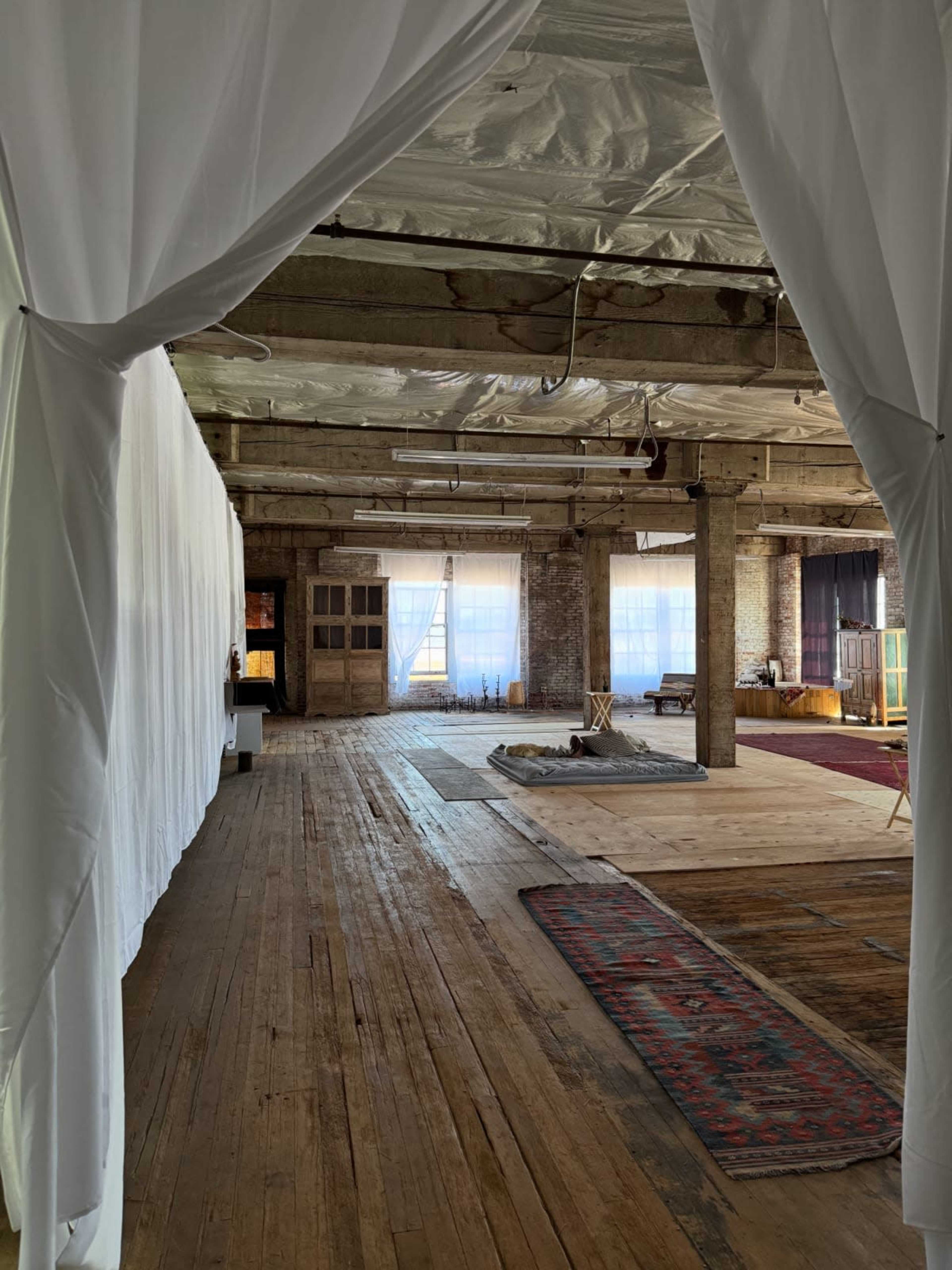 The image shows a spacious, rustic interior with wooden floors, exposed beams, and soft fabric drapes framing the entrance to the room.