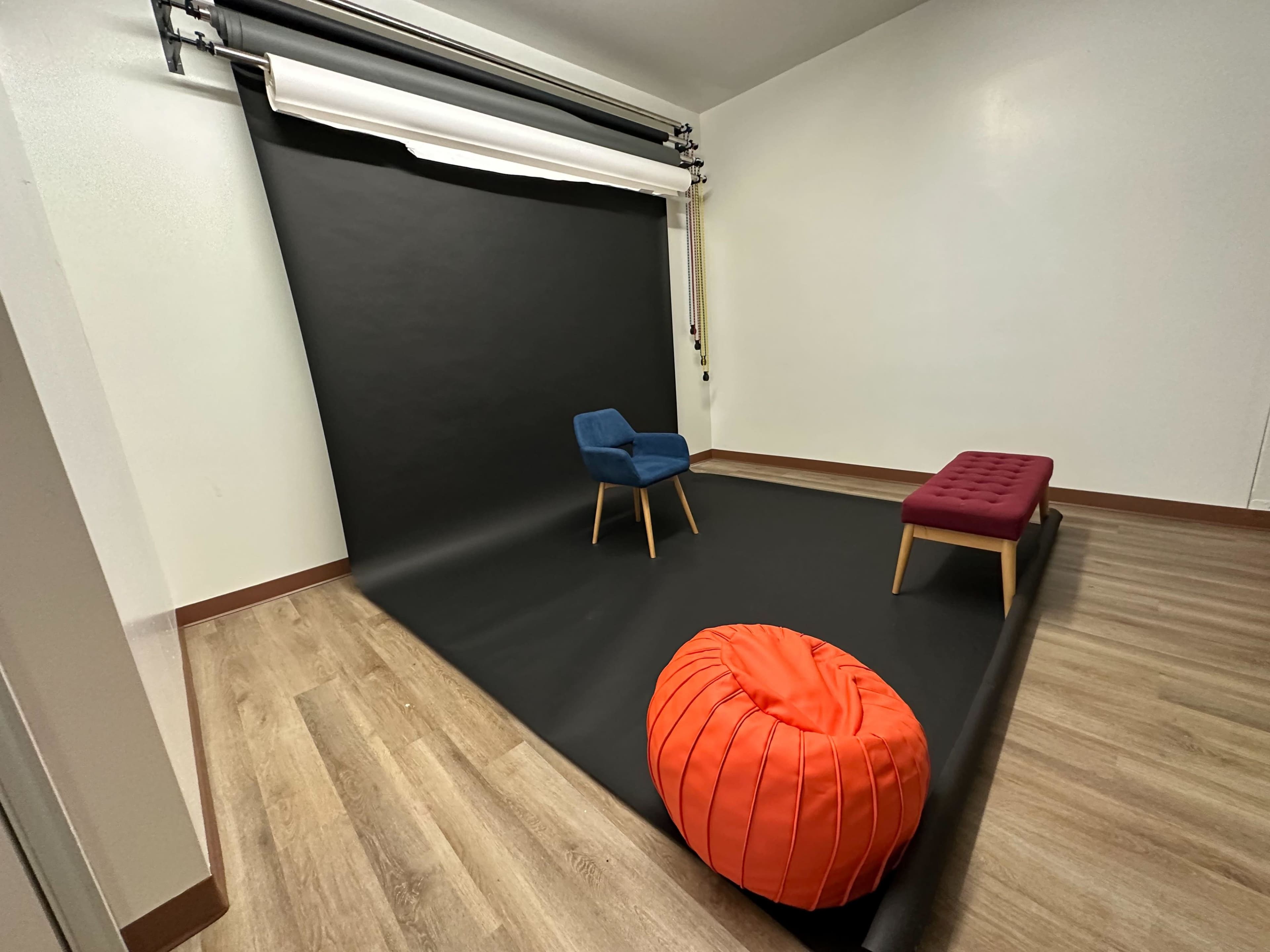 The image shows a photography studio setup with a black backdrop, a blue chair, a red ottoman, and an orange beanbag on a wooden floor.