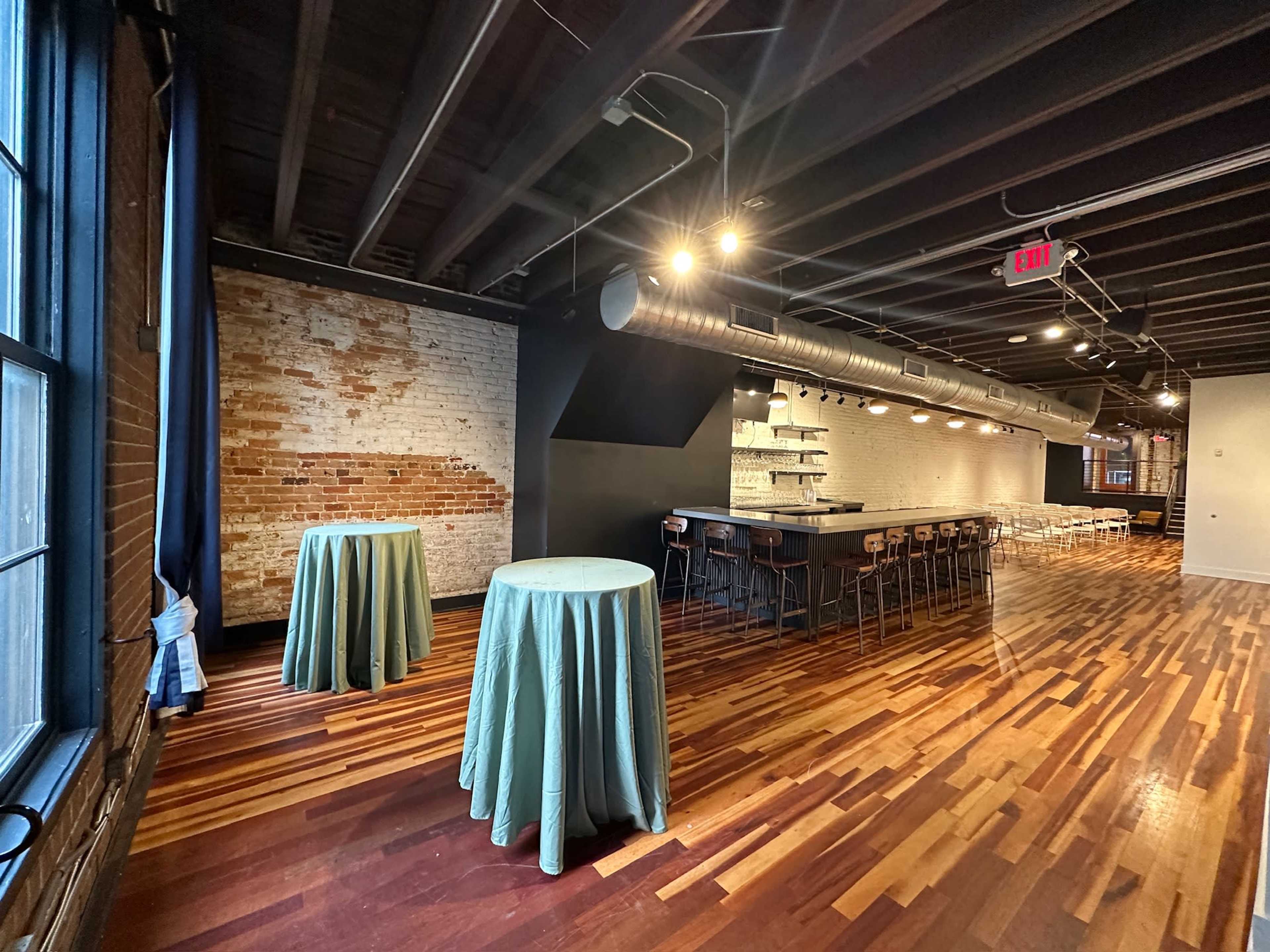 The interior of a modern venue features two round tables with light blue tablecloths, a long bar area, and a combination of exposed brick and wooden flooring.
