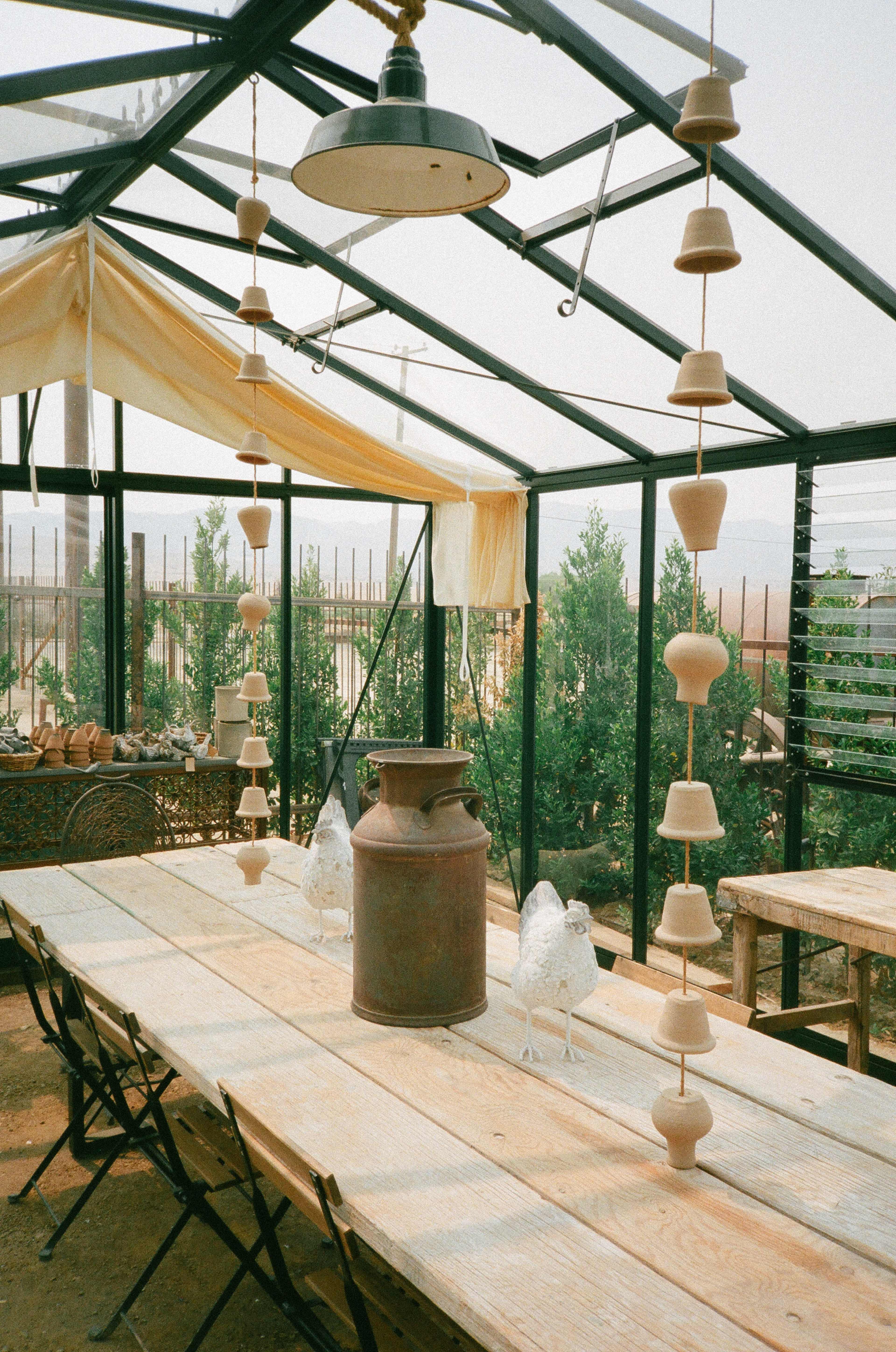 A greenhouse interior with a long wooden table, decorative pottery hanging from the ceiling, and a vintage milk can as a centerpiece.