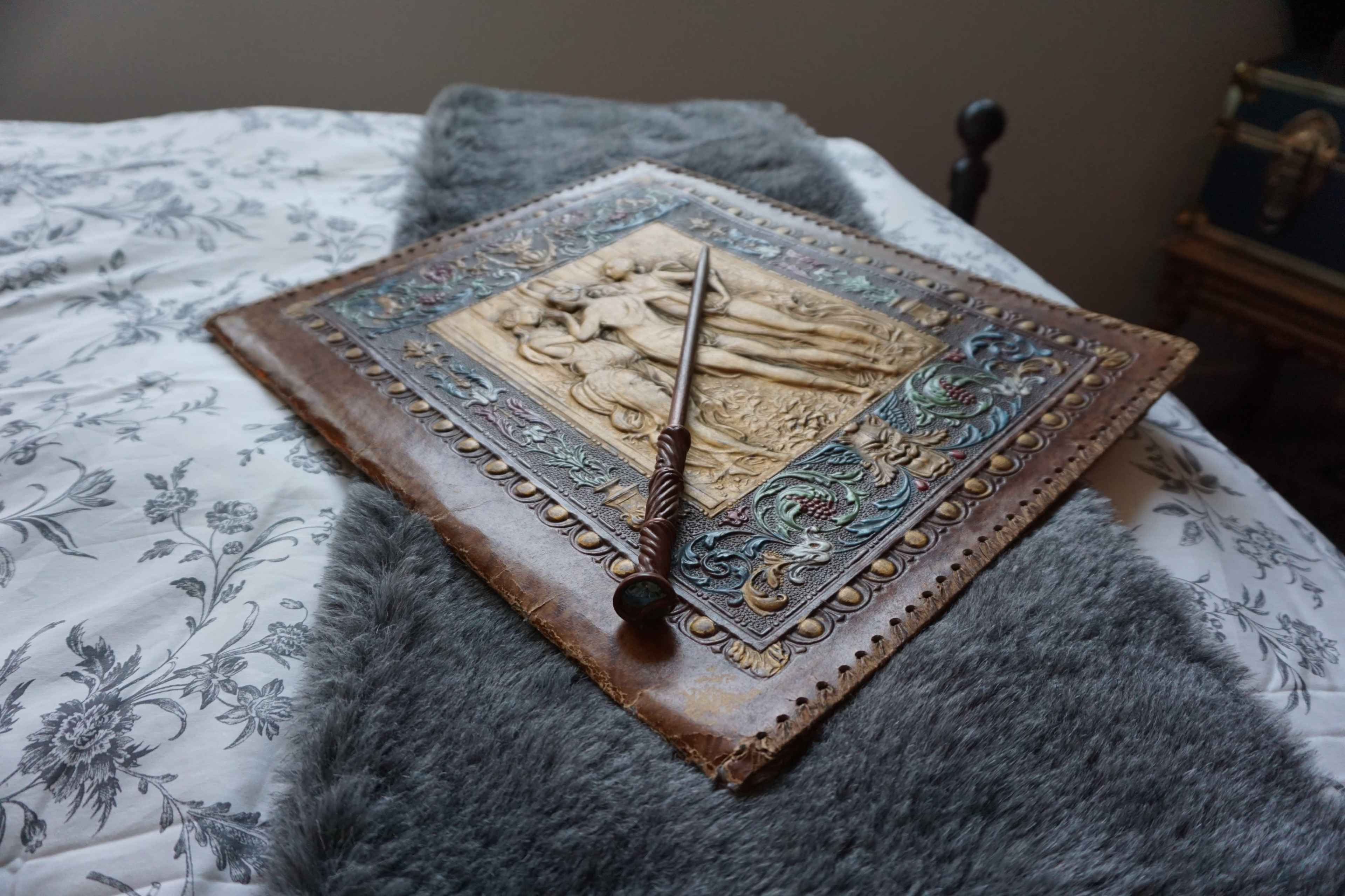 A textured leather-bound book with an embossed cover lies on a gray fur throw on a bed with floral-patterned sheets.