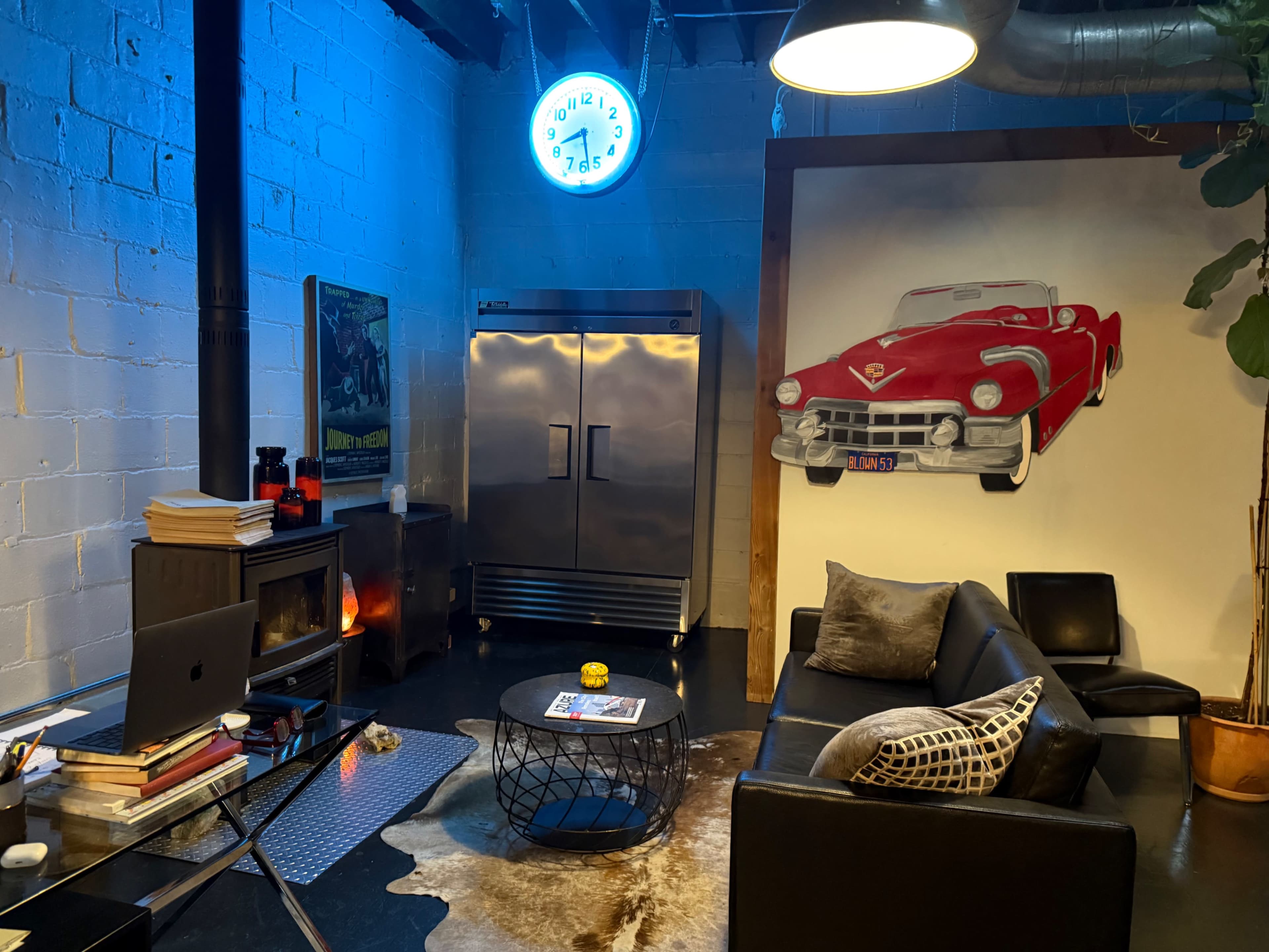 The image shows a modern lounge area with a black leather sofa, a round coffee table, a wall-mounted clock, a stainless steel refrigerator, and a large wall art featuring a red classic car.