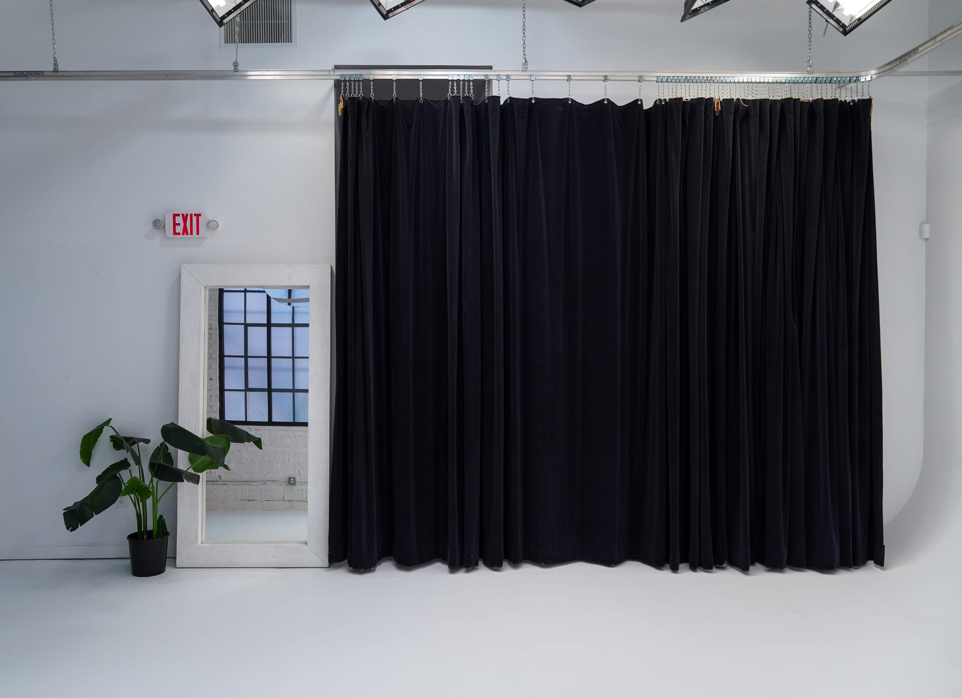 A spacious studio features a large black curtain, a mirror, and a potted plant against a minimalist white backdrop.
