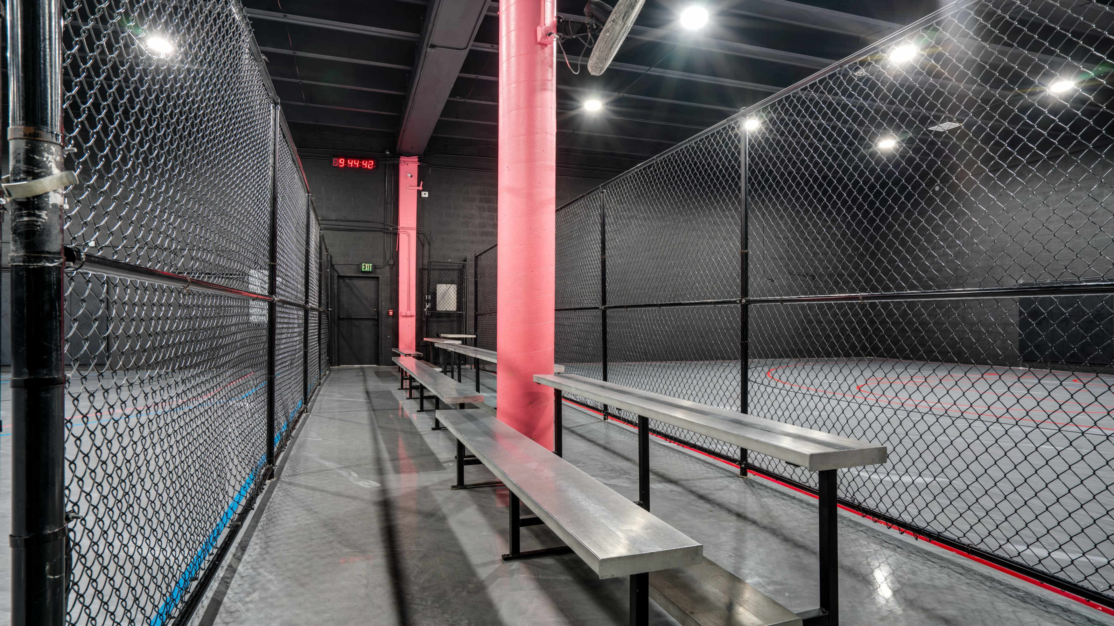 The image shows a fenced indoor sports facility with metal benches along the sides and a basketball court in the background.