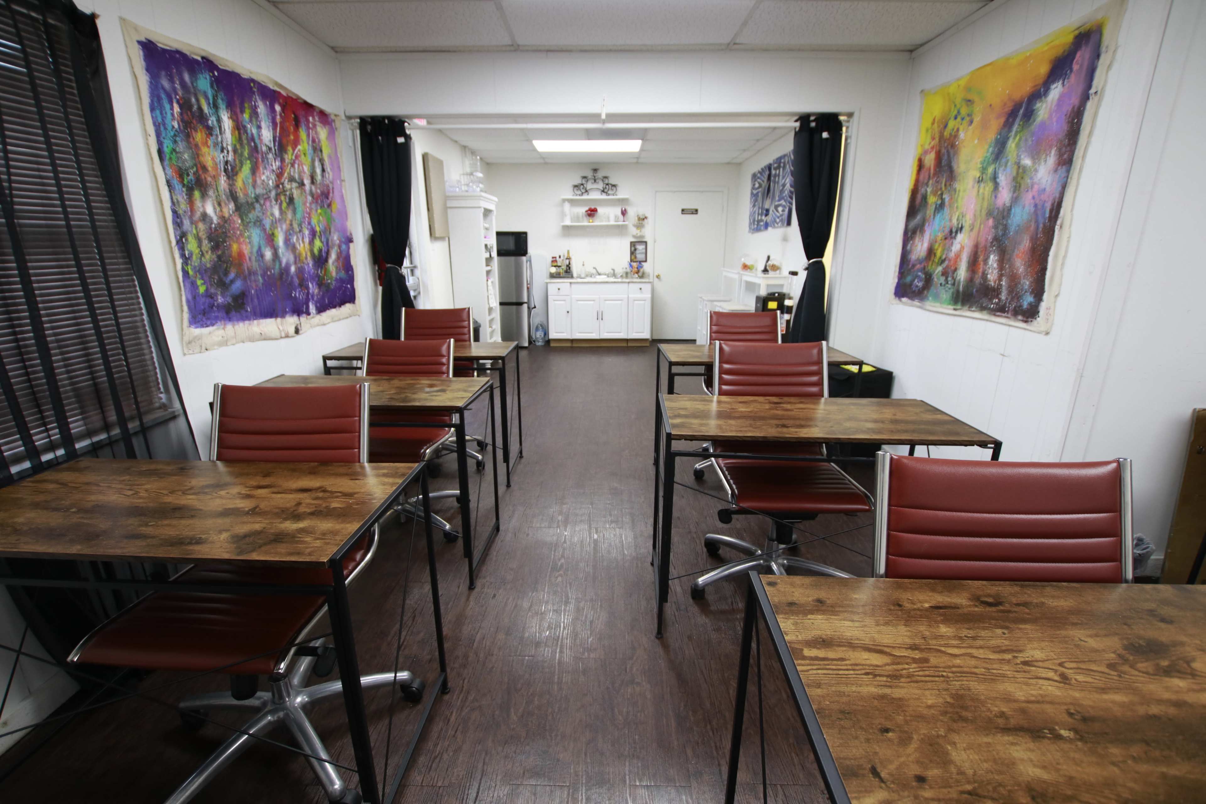 The image depicts a spacious room with several wooden tables and chairs arranged in two rows, leading to a kitchenette area at the back, adorned with colorful artwork on the walls.