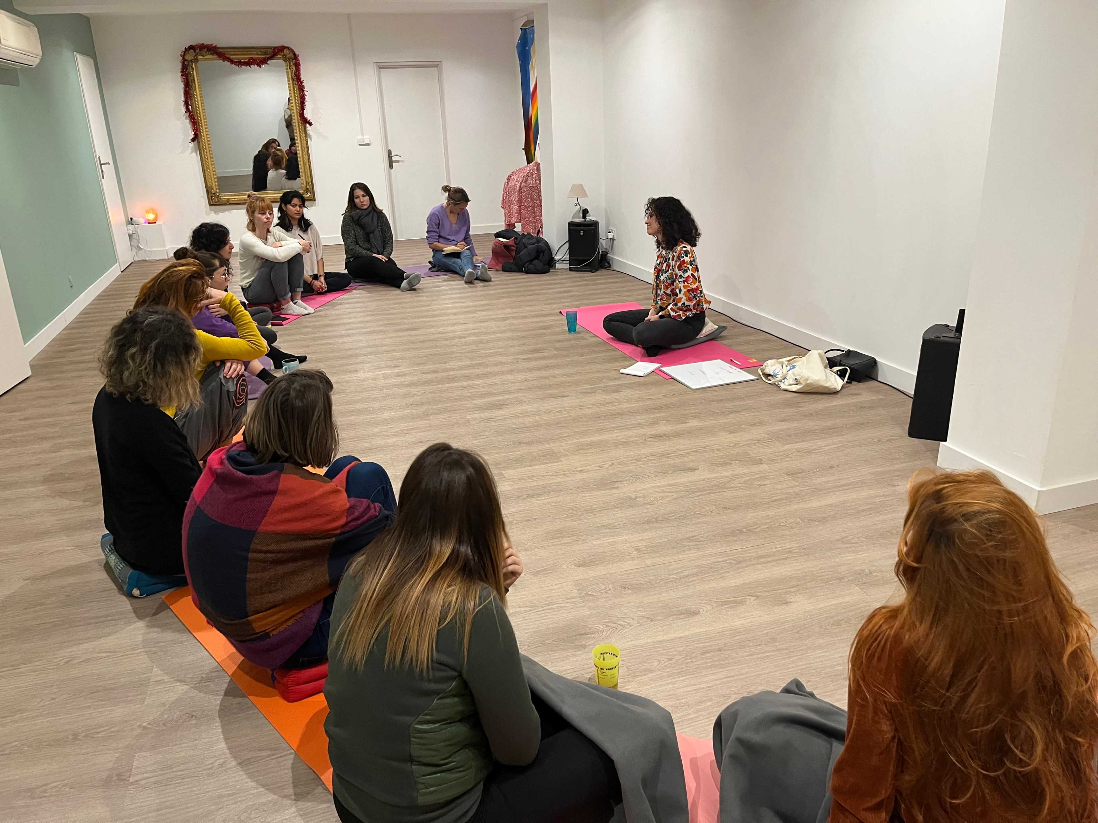 A group of people sits on mats in a spacious room, attentively listening to a speaker who is seated at the front.