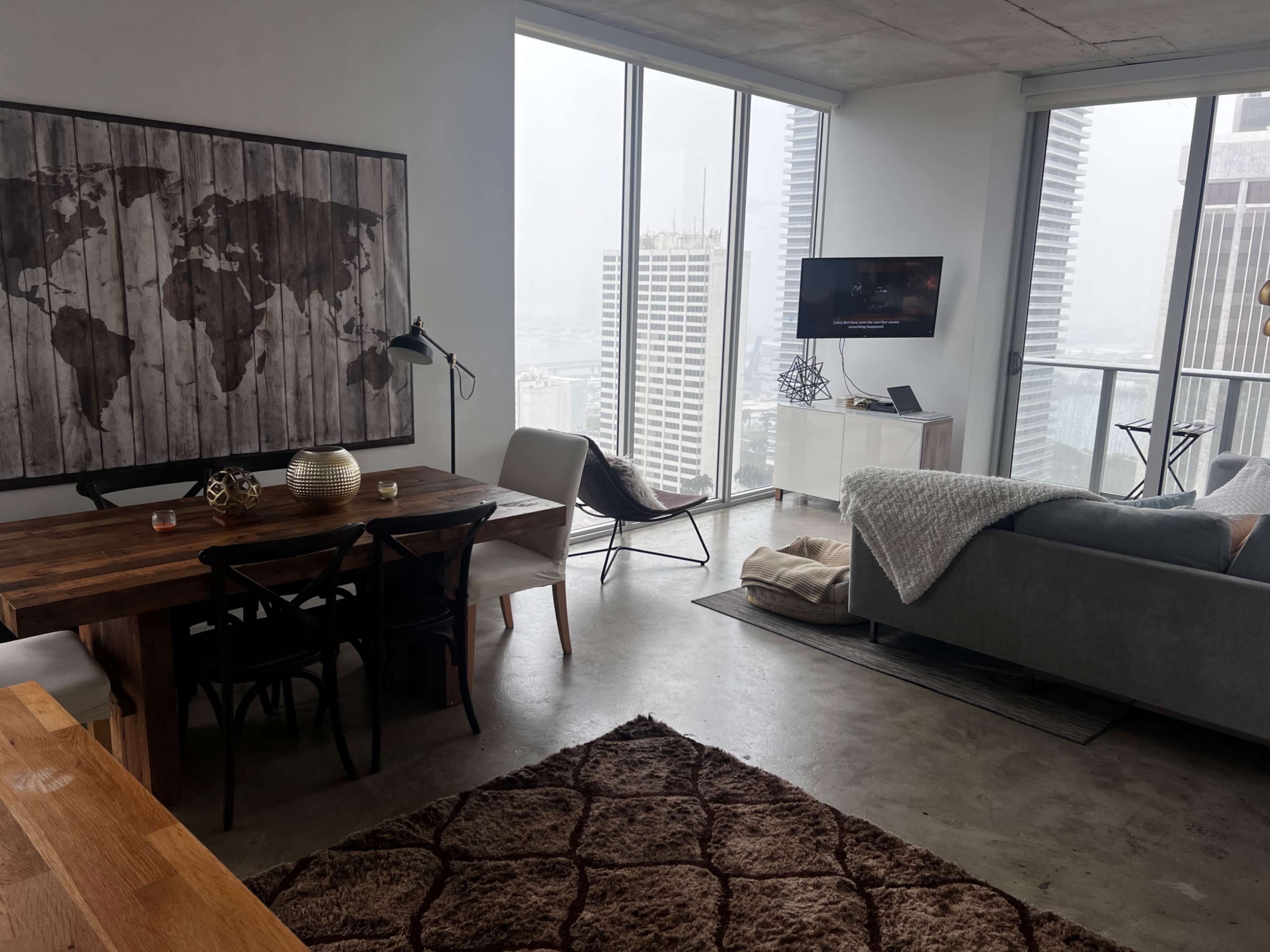 The image shows a modern apartment with a dining area, a living space featuring a sofa and TV, and large windows displaying a city view.