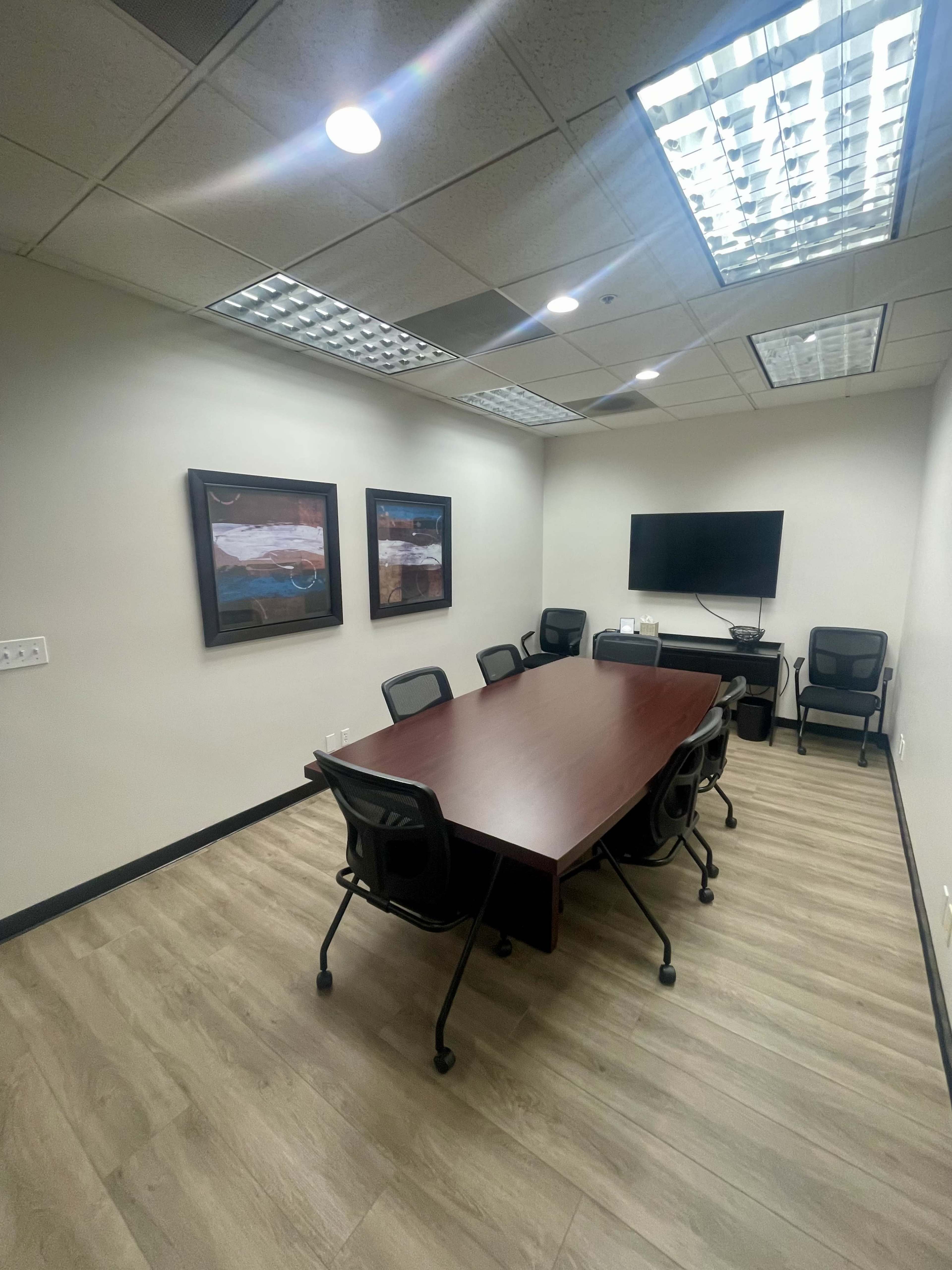A modern conference room with a long wooden table surrounded by black chairs, two abstract paintings on the walls, and a wall-mounted television.