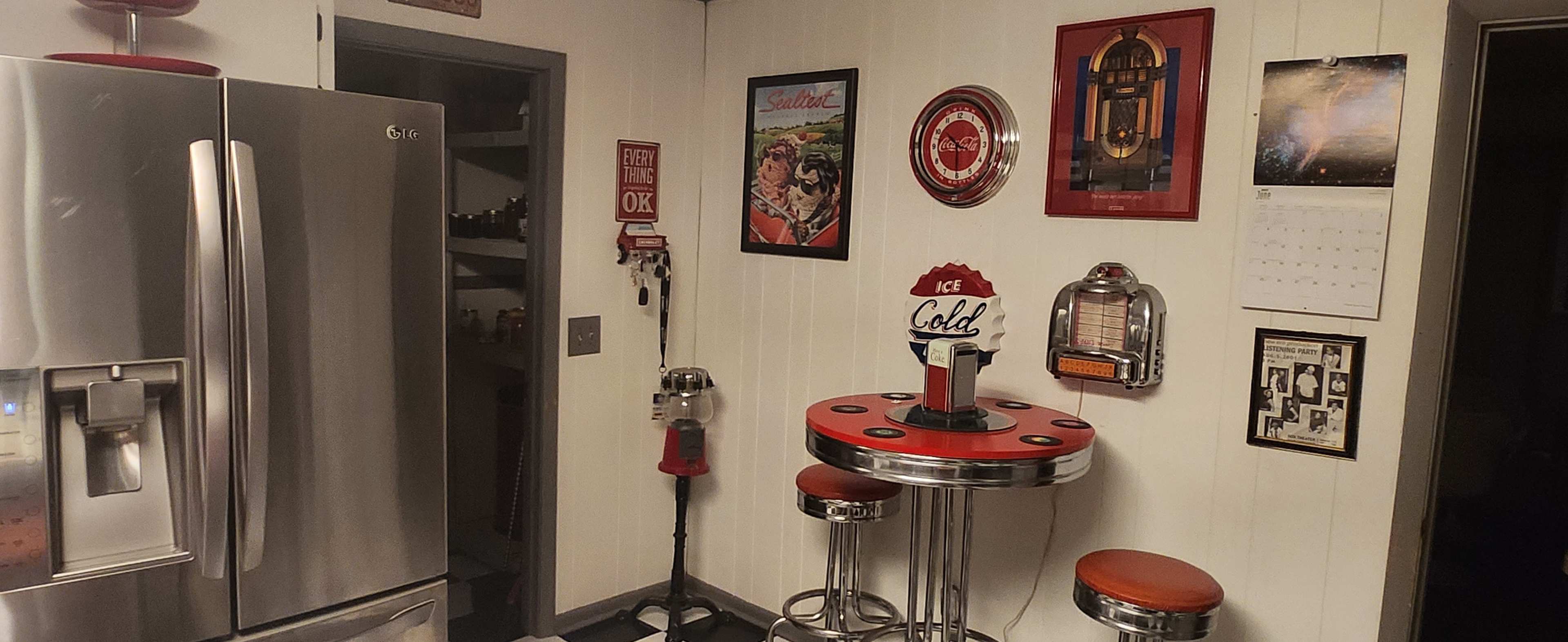 The image shows a retro-style kitchenette featuring a round red and silver table with two stools, a vintage jukebox decor, and various framed wall art.