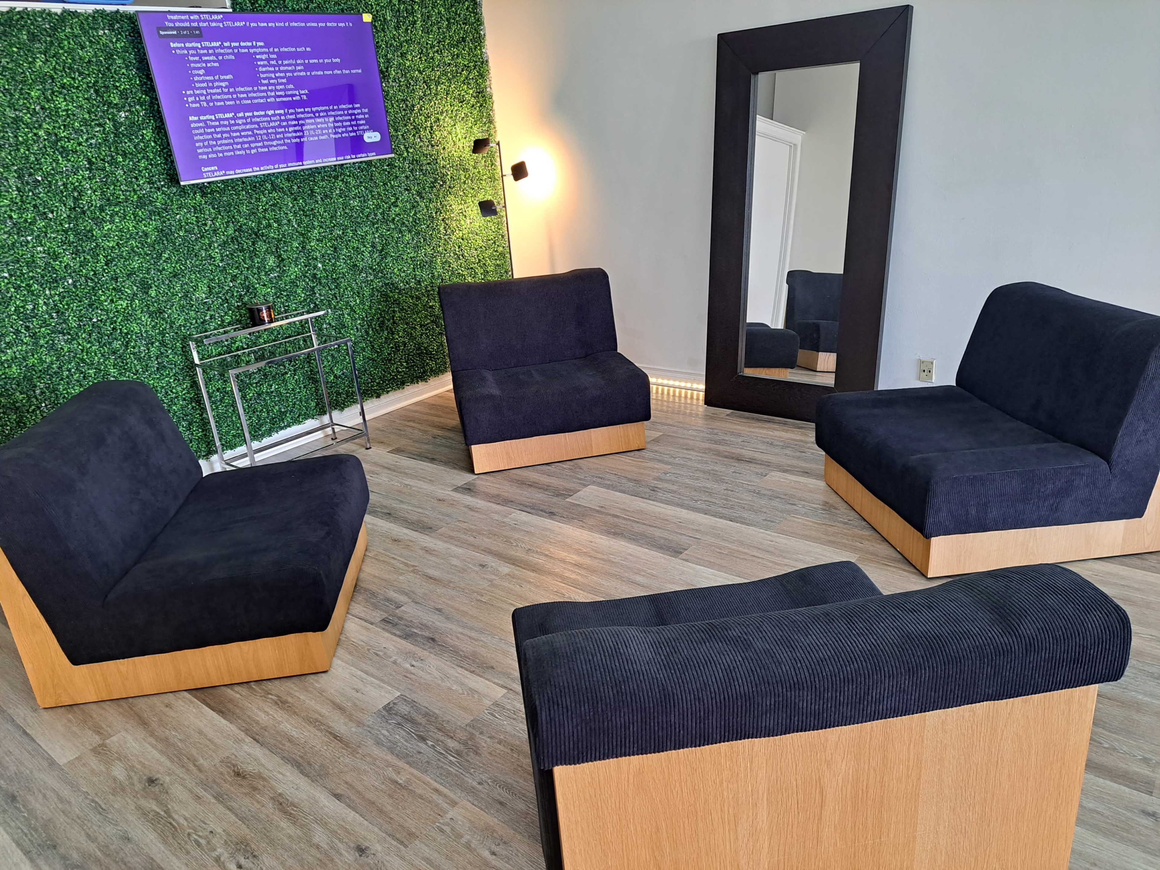 A modern lounge area with four upholstered seating units arranged around a small table, a large mirror, and a wall covered in greenery, while a screen displays information.