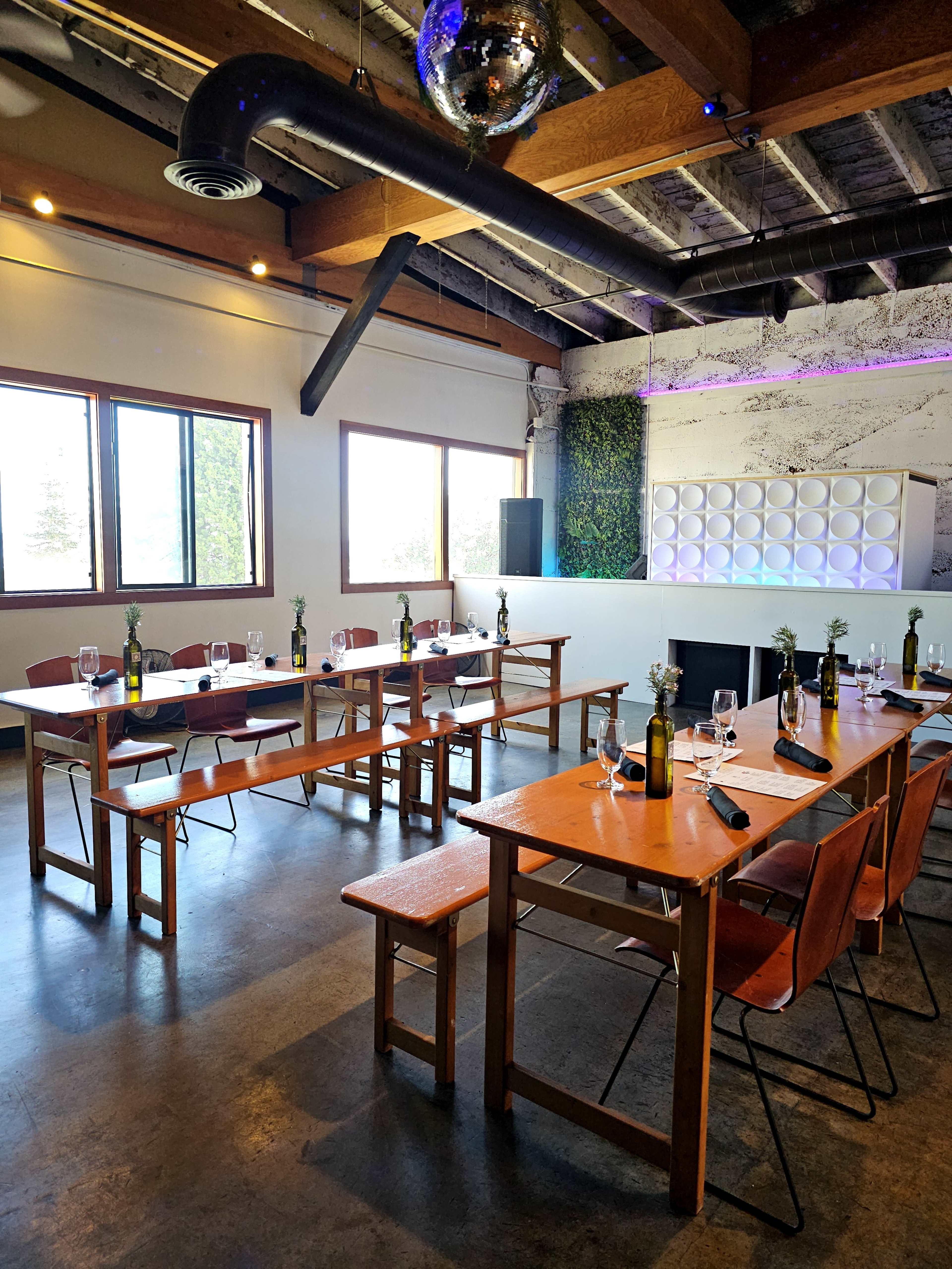 The image shows a modern dining room setup with two long tables arranged for a meal, featuring empty wine bottles, glasses, and place settings, under exposed beams and a disco ball.