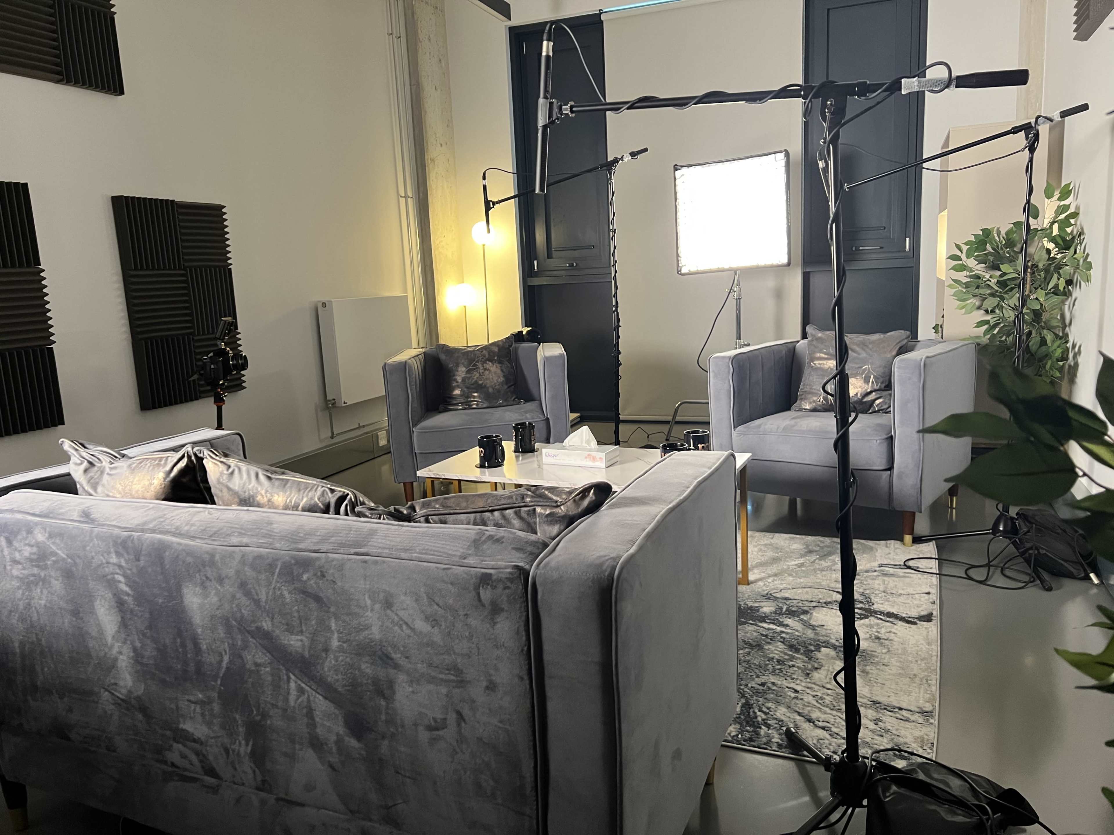 A modern, well-lit interview setup with two gray sofas, a coffee table, and microphone stands in a studio environment.