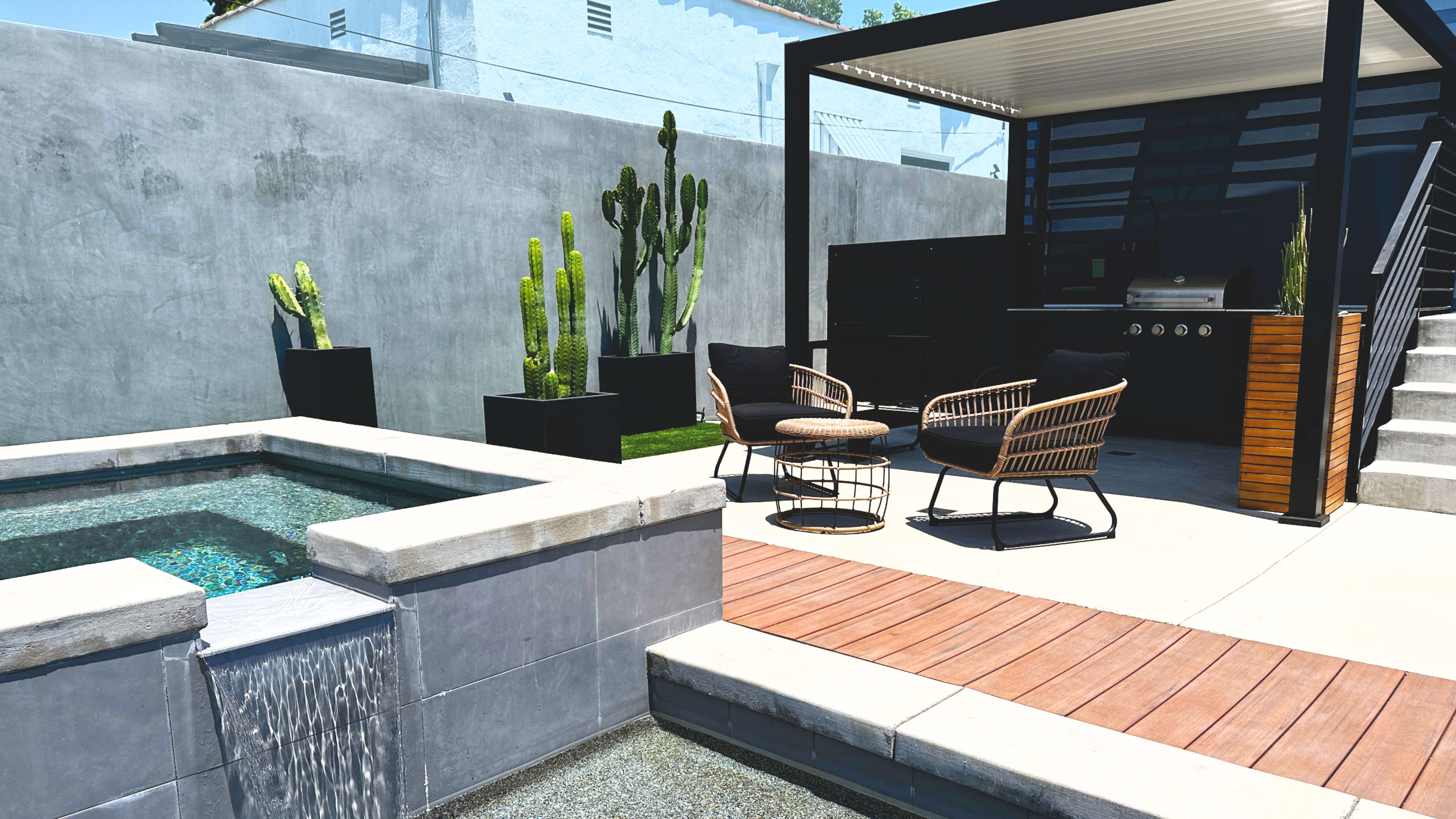 A modern backyard with a seating area beside a small pool, featuring cacti in planters and a barbecue grill under a shaded structure.