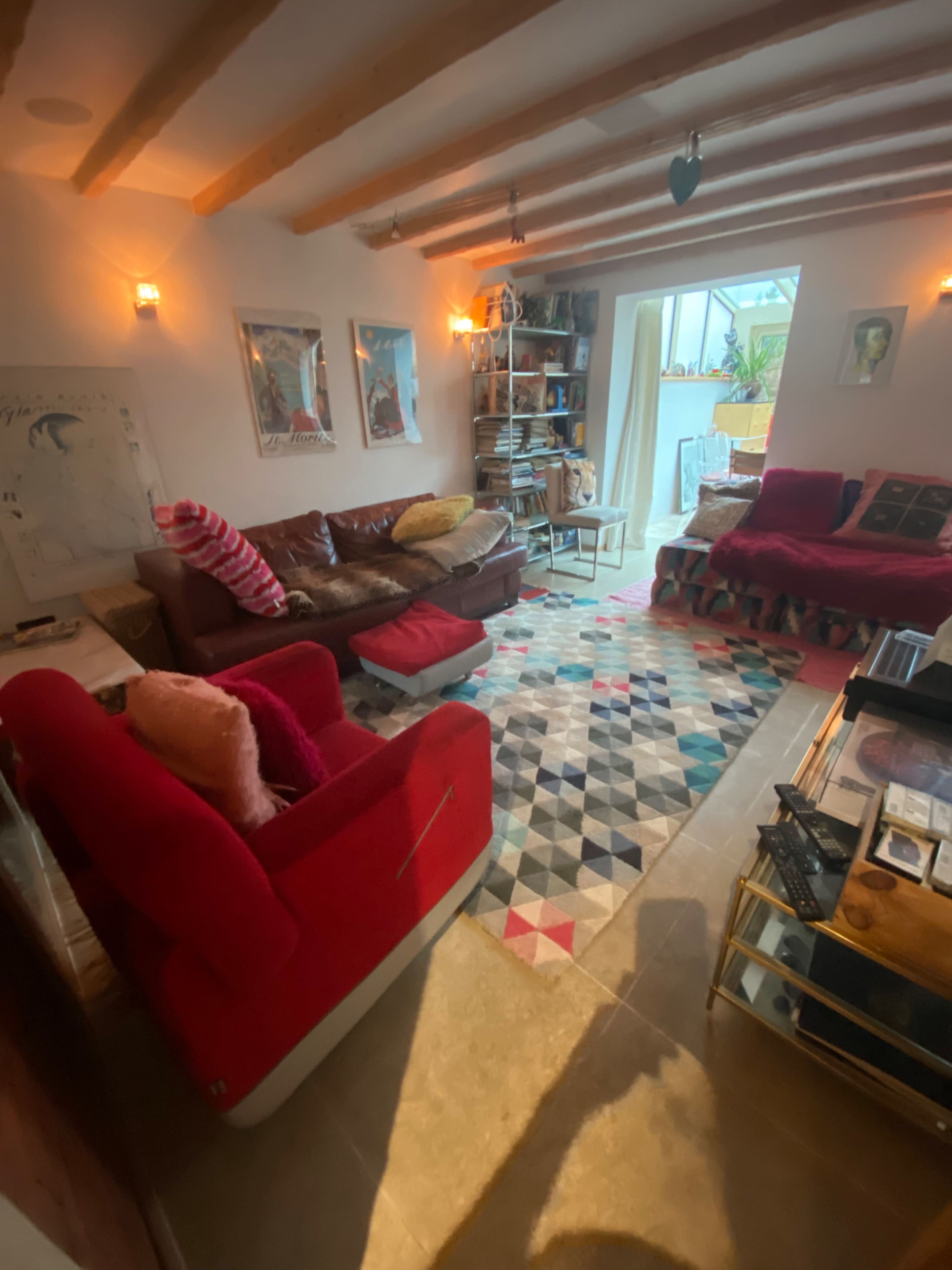 A cozy living room features a mix of red and brown furniture, a patterned rug, and bookshelves lined with items, illuminated by natural light from a nearby doorway.