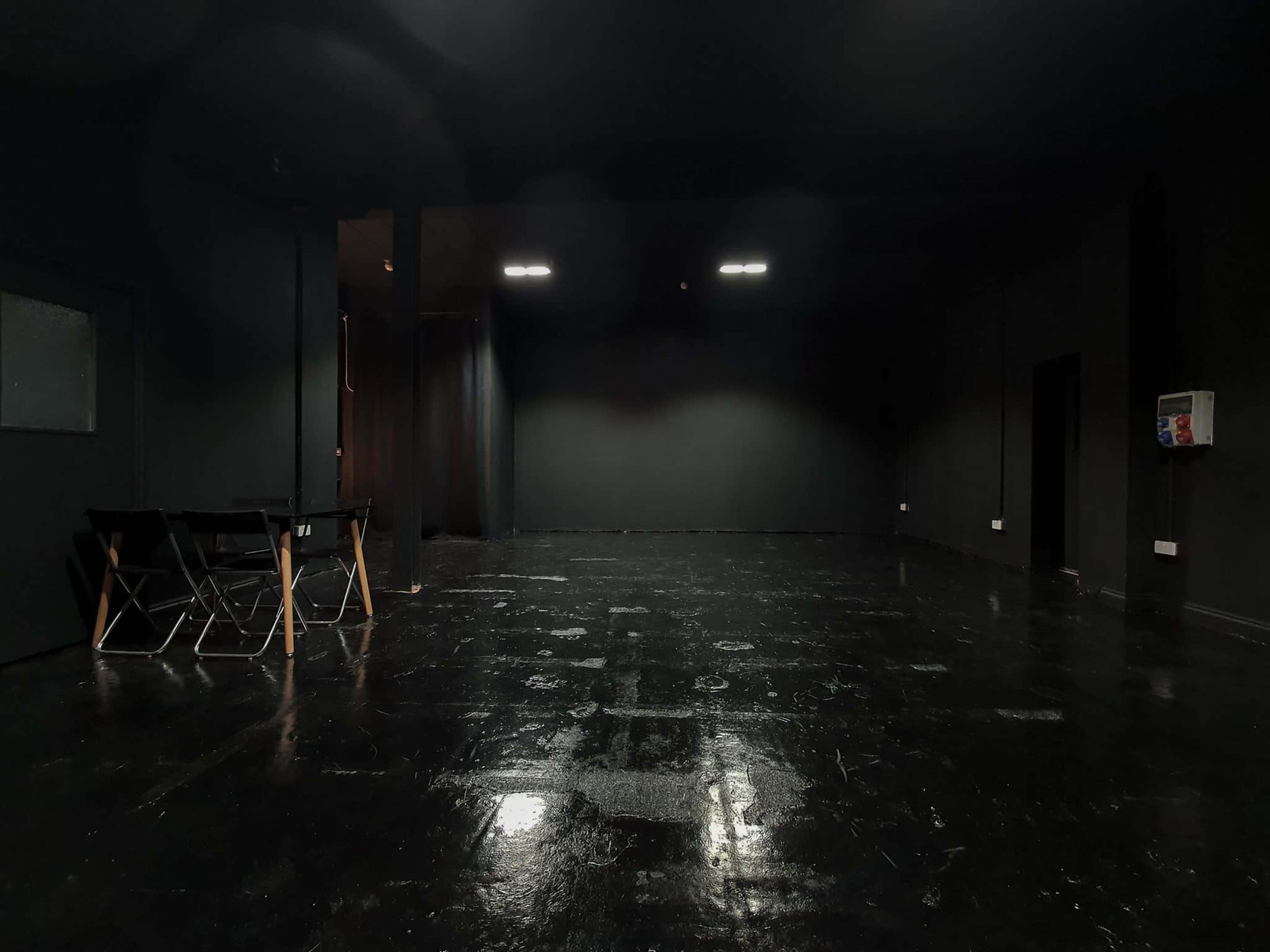 A dimly lit, empty room with black walls and a glossy black floor, featuring a table and chairs on one side.