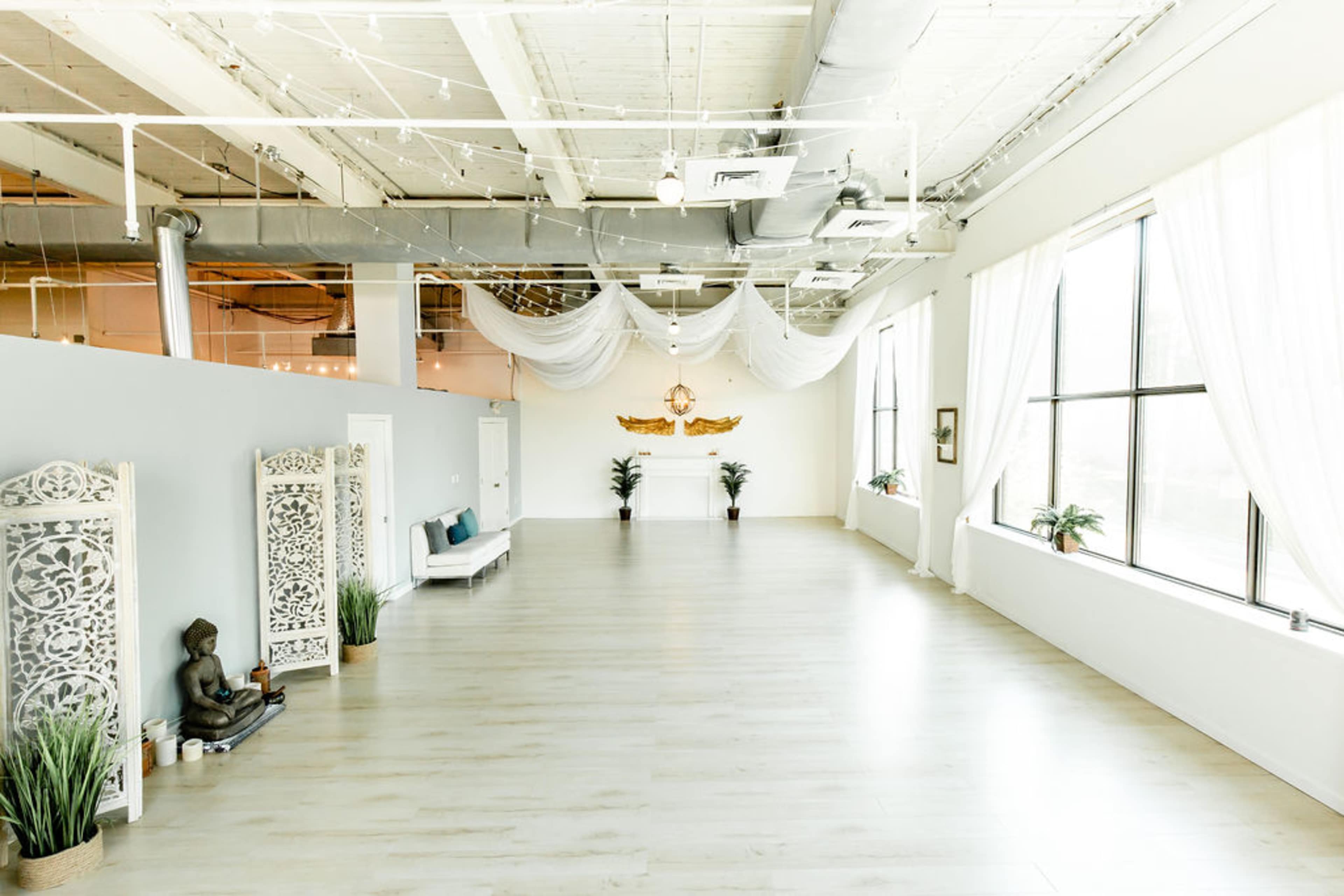 The spacious interior features light-colored wooden flooring, large windows with sheer curtains, and minimal decor including a Buddha statue and ornamental screens.
