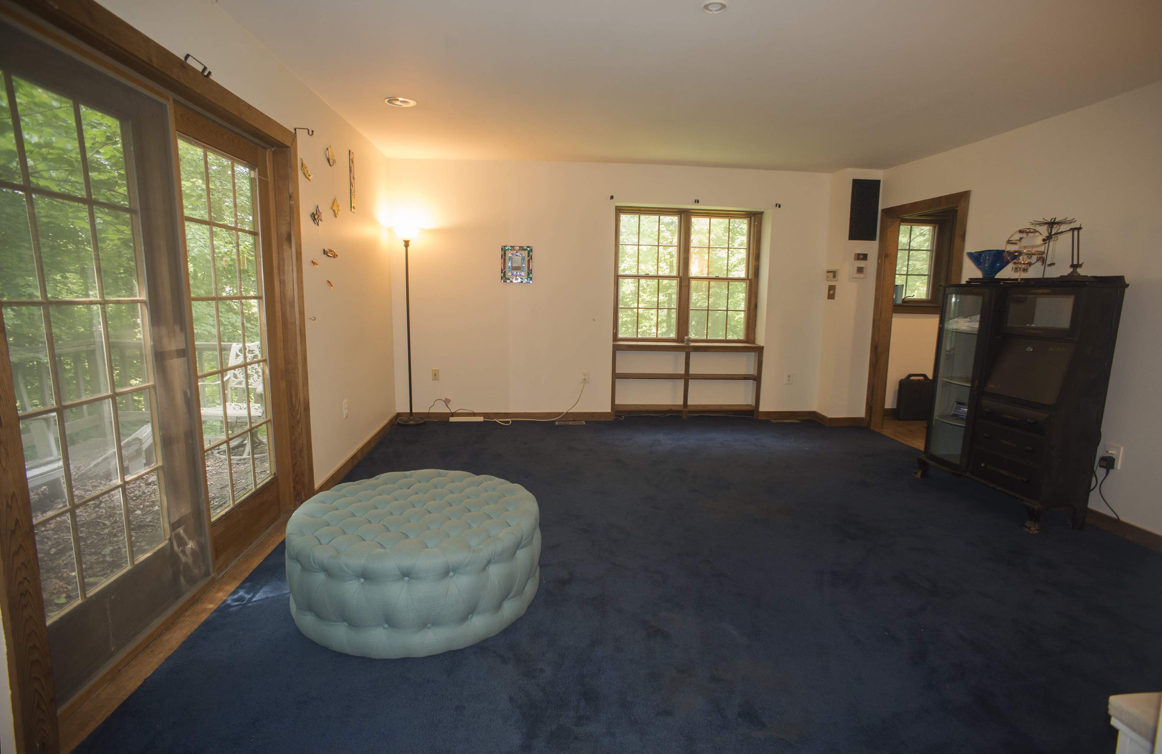 The room features a blue carpet, large windows with natural light, a round tufted ottoman, and a wooden shelf against the wall.