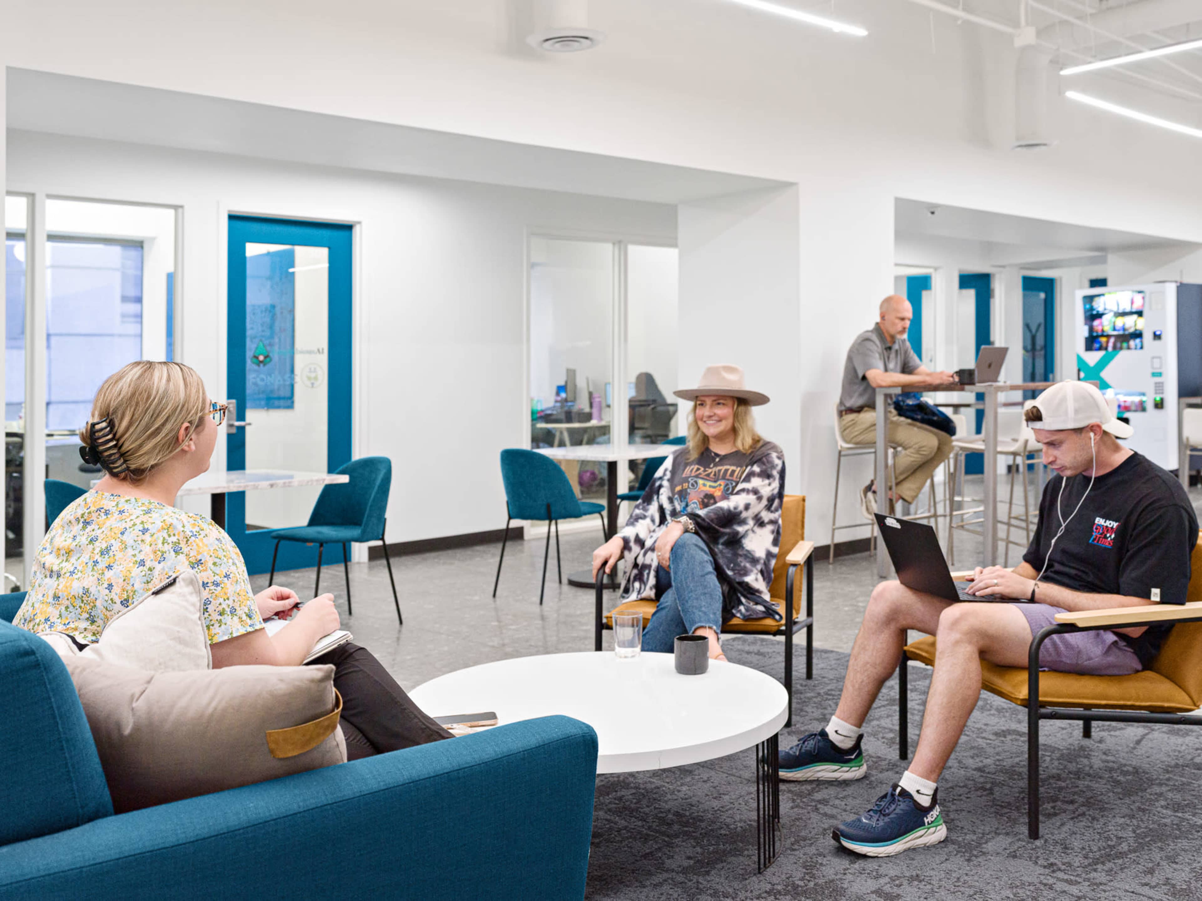 Three people are engaged in different activities in a modern, brightly lit lounge area with blue chairs and a communal workspace in the background.
