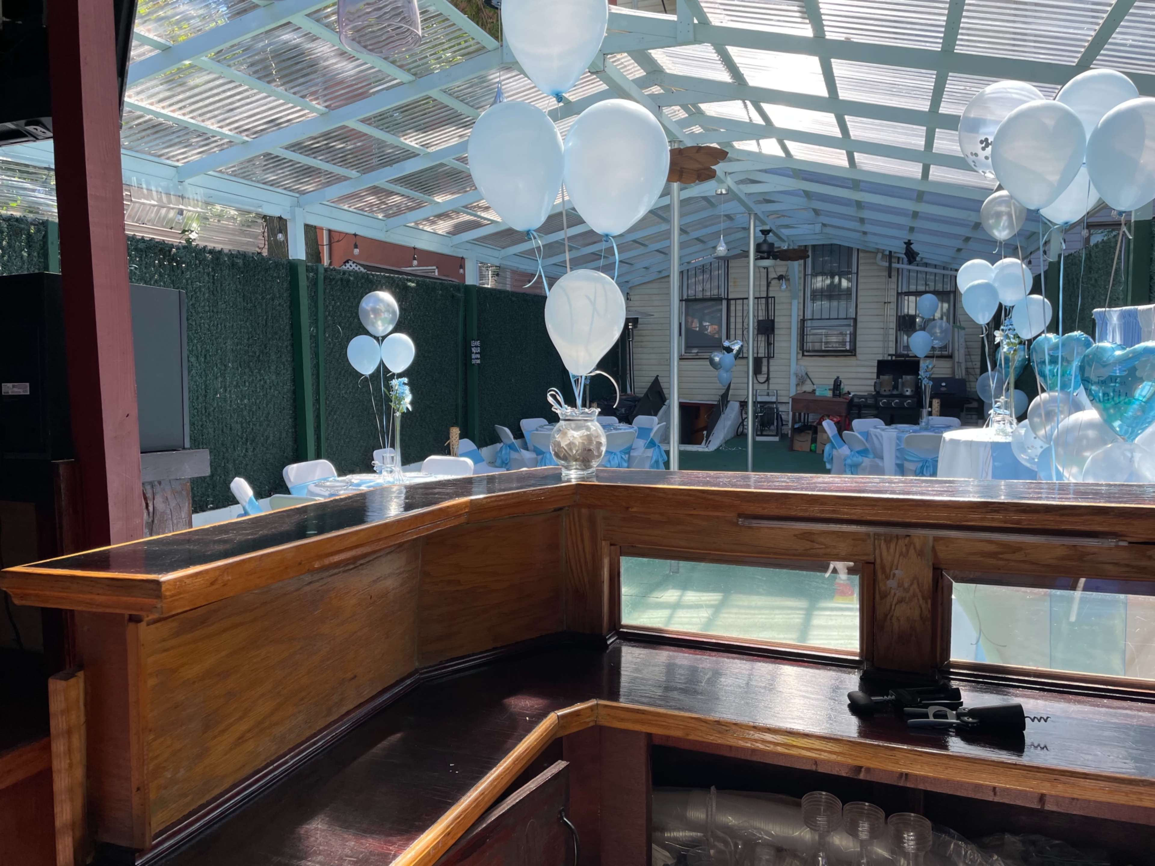The scene shows a decorated outdoor venue with blue balloons and tables set up for an event, viewed from a wooden counter.