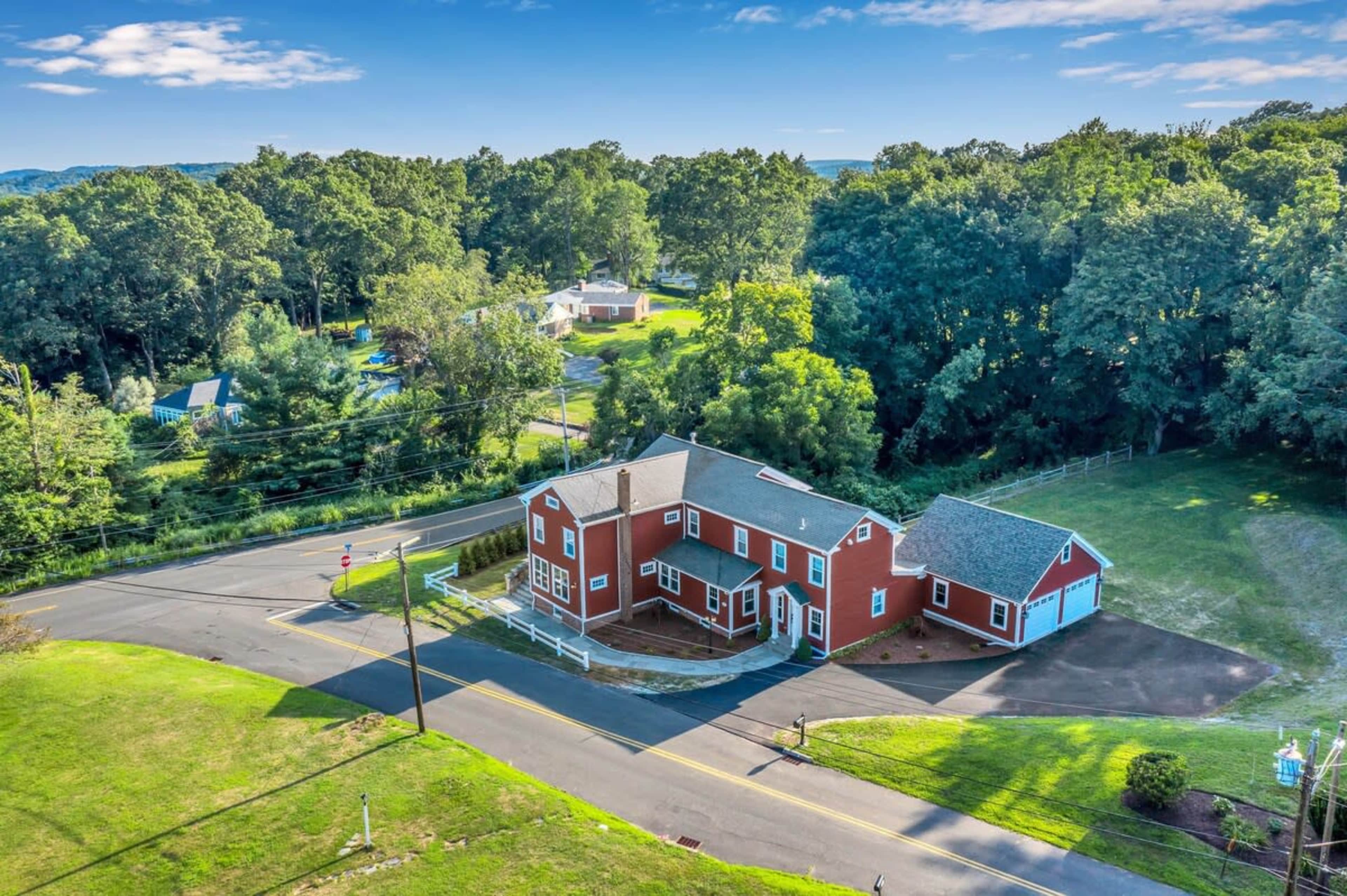 A red two-story house at the intersection of two roads, surrounded by trees and grassy areas.