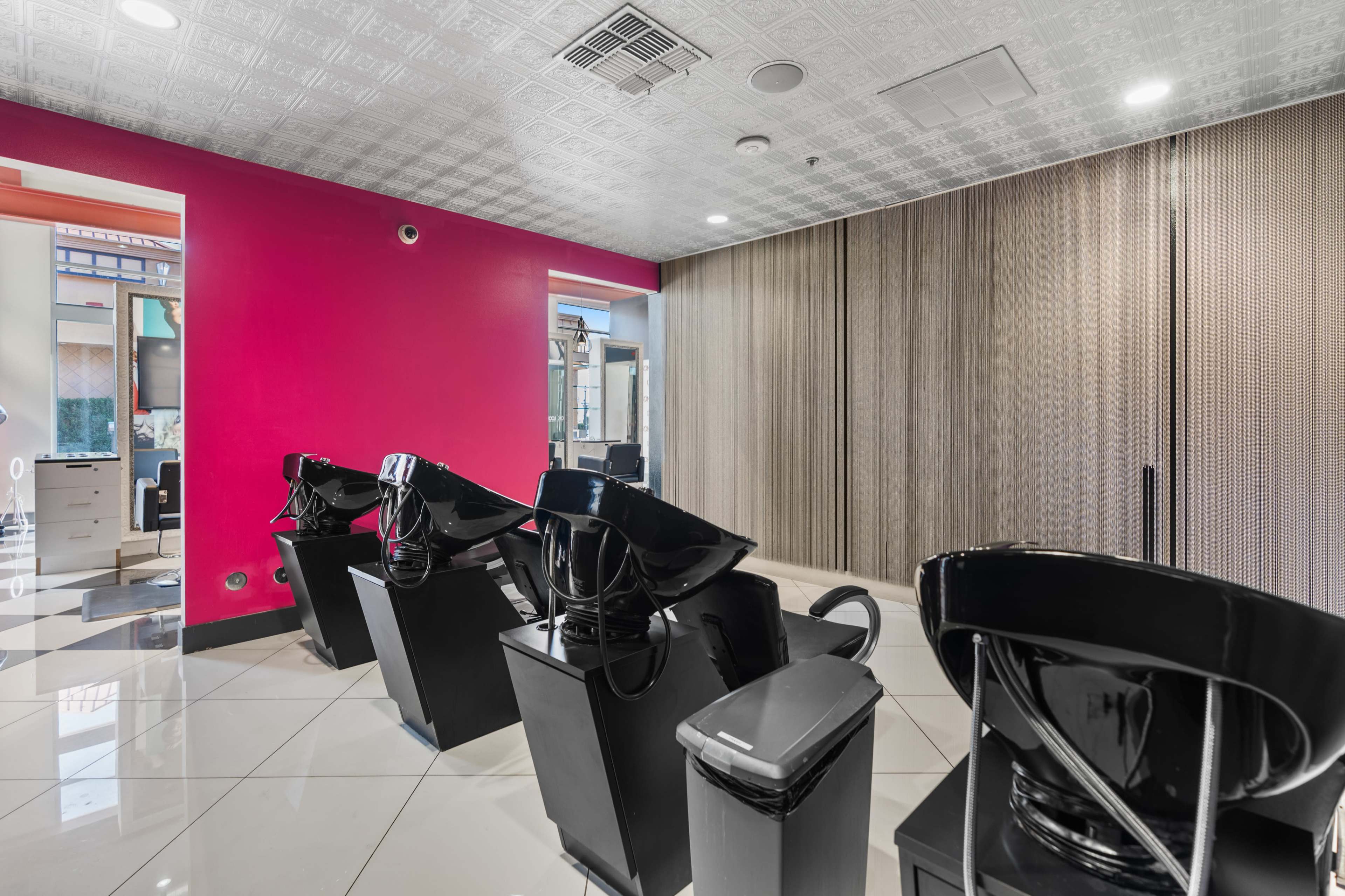 A modern hair salon with black shampoo stations, white tile flooring, and a bright pink accent wall.