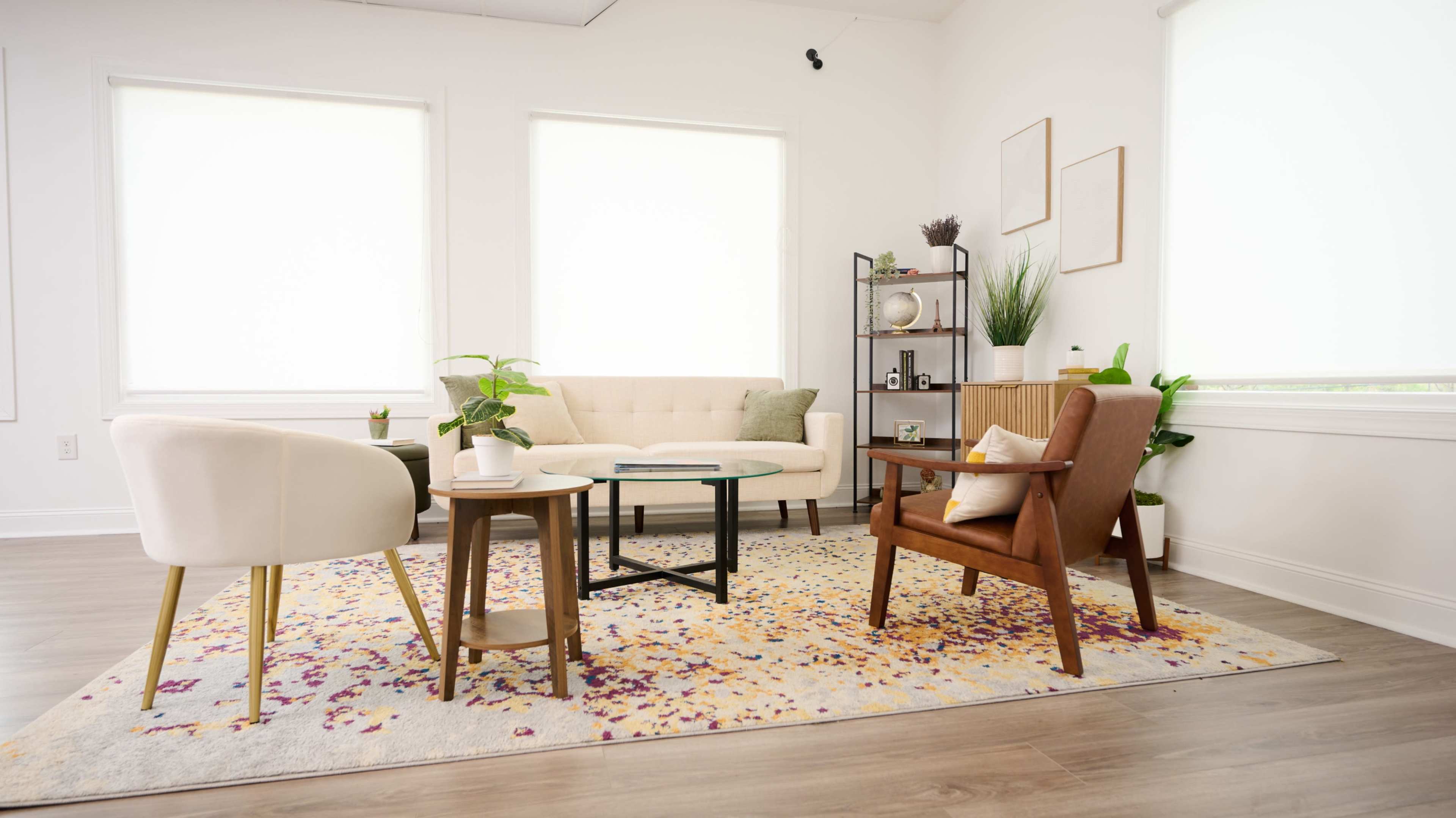 A bright and modern living room features a beige tufted sofa, two accent chairs, a coffee table, and decorative plants, all arranged on a colorful area rug.