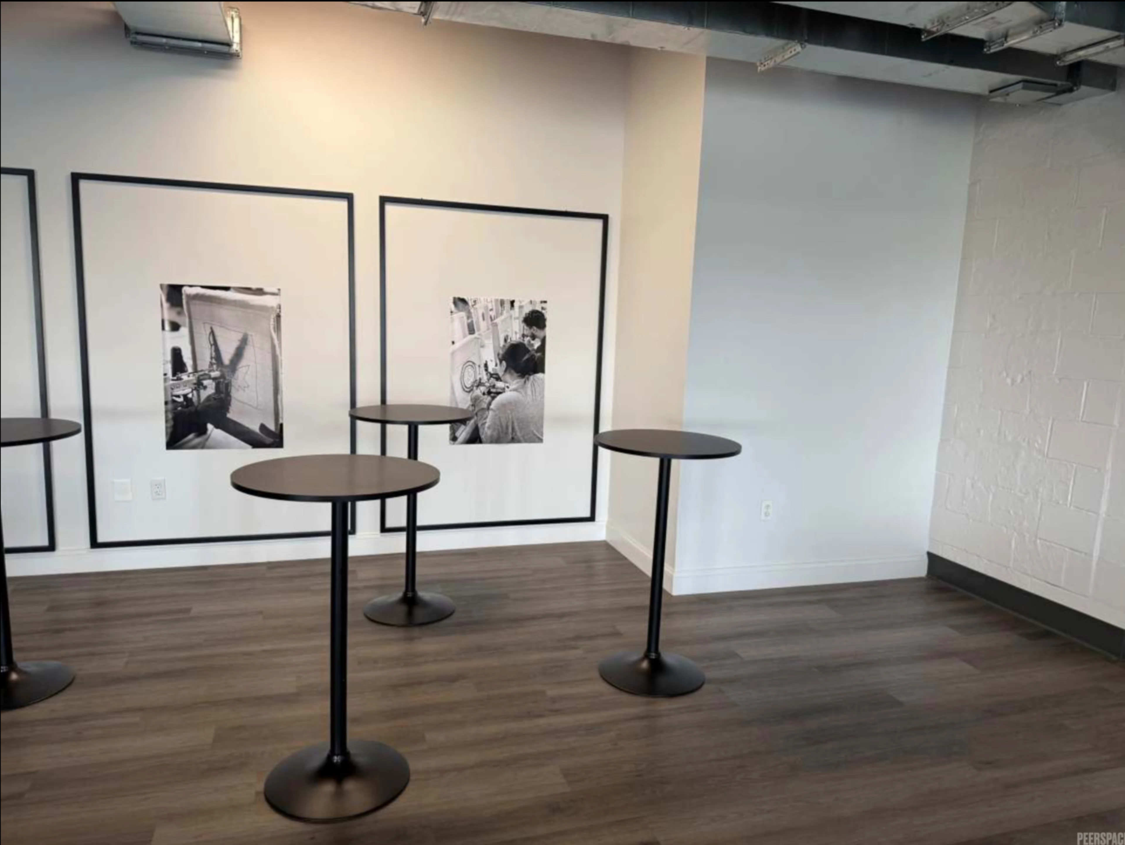 The image shows a minimalist room with three tall black tables and two large framed black-and-white photographs on the wall.