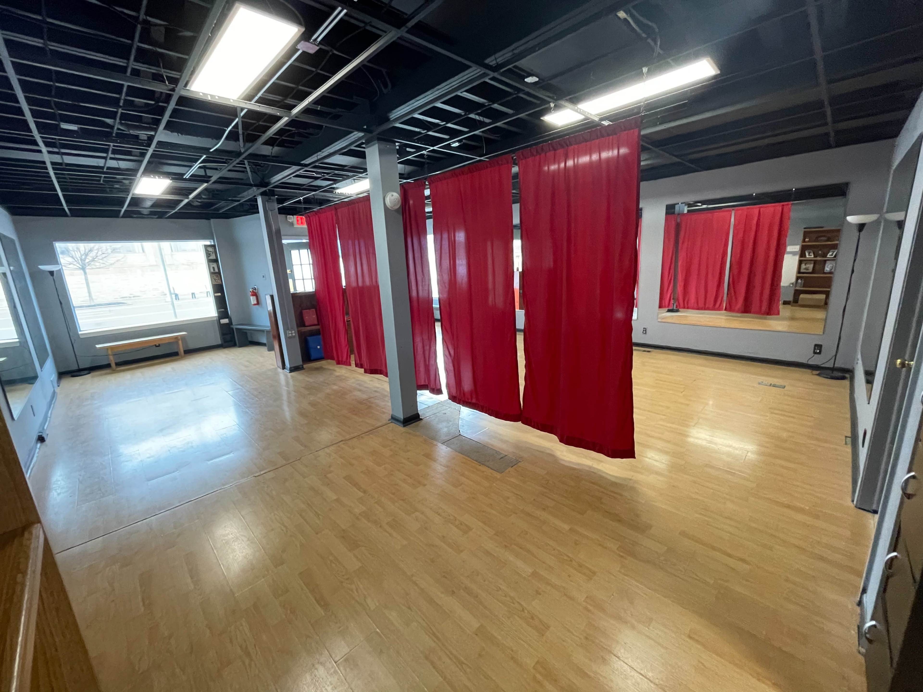 A spacious room with wooden flooring, large windows, and red curtains dividing the space, along with a mirror on one wall.