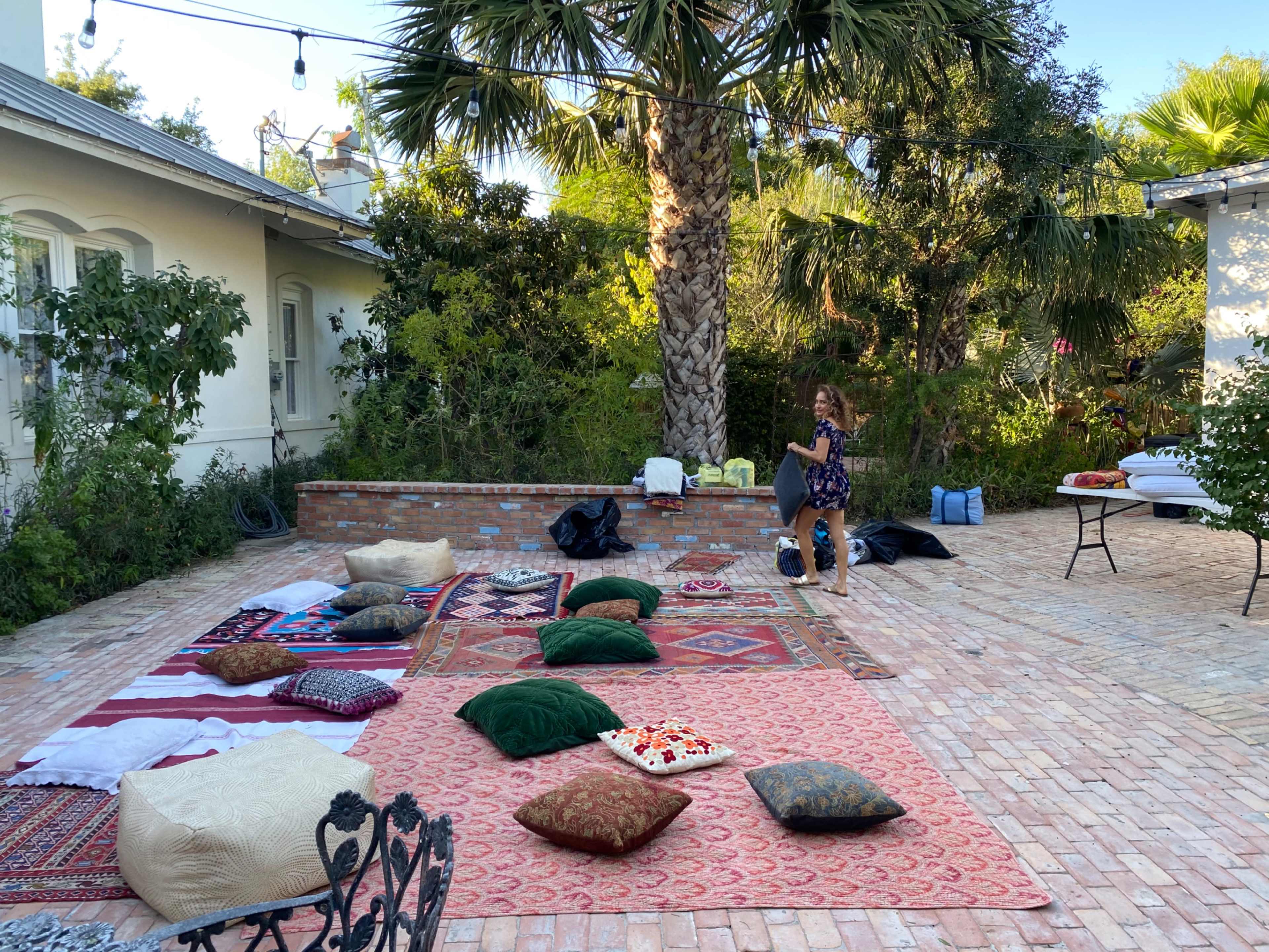 A woman sits among colorful rugs and cushions arranged on a brick patio surrounded by greenery and palm trees.