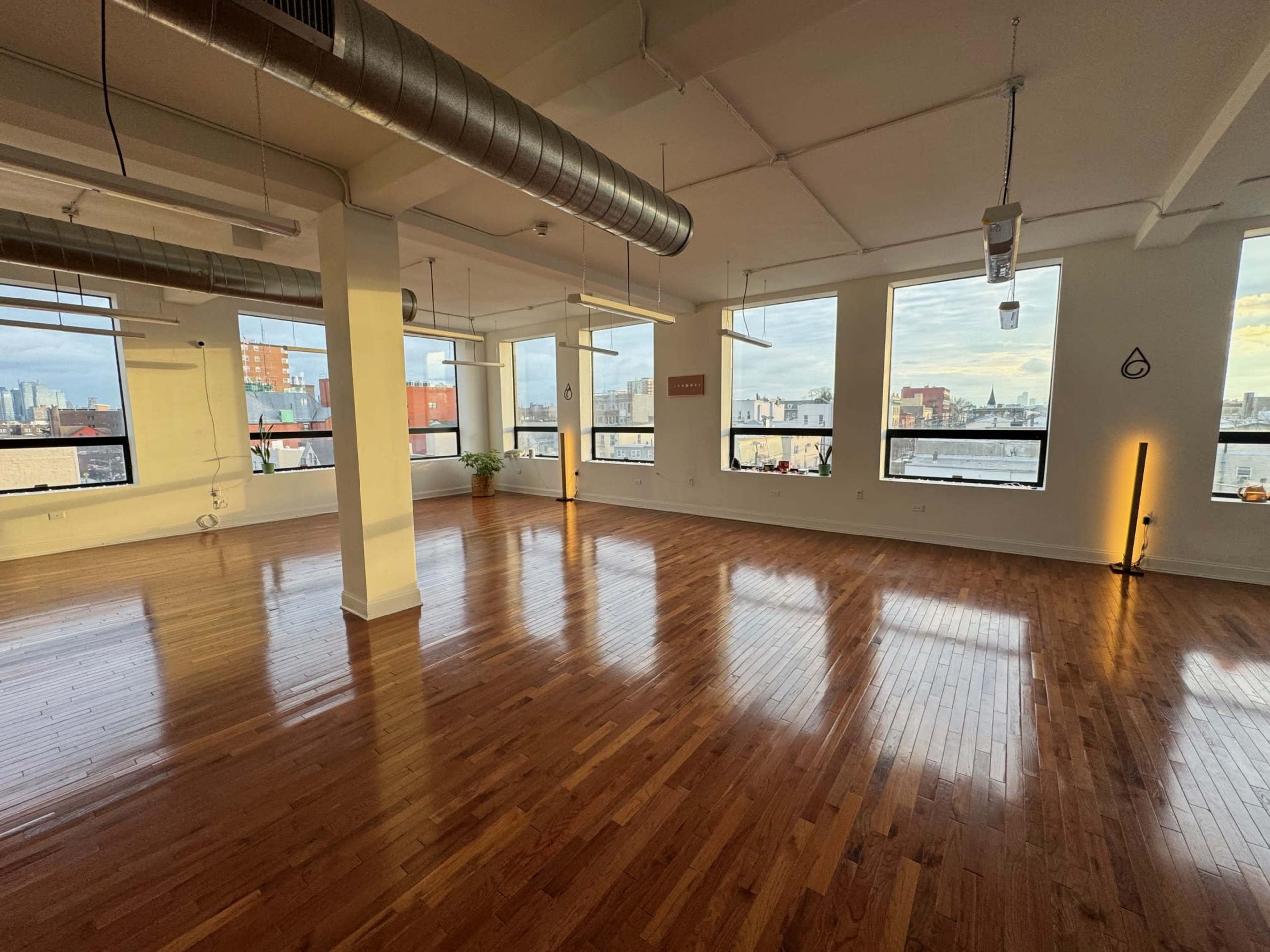 The image shows a spacious, empty room with hardwood floors and large windows allowing natural light to enter.