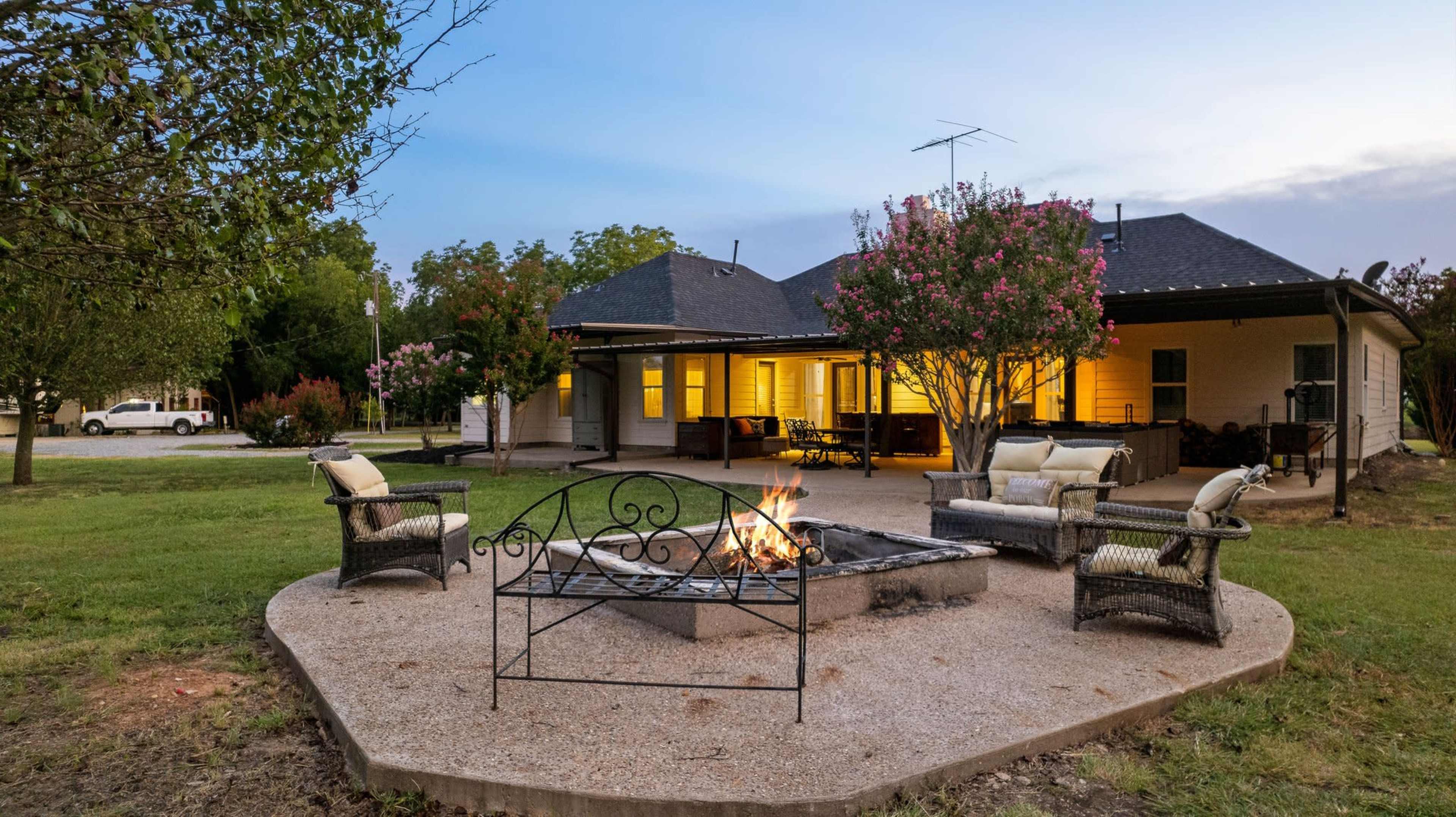 A fire pit surrounded by seating is set in a grassy yard beside a house with lights glowing from the windows.