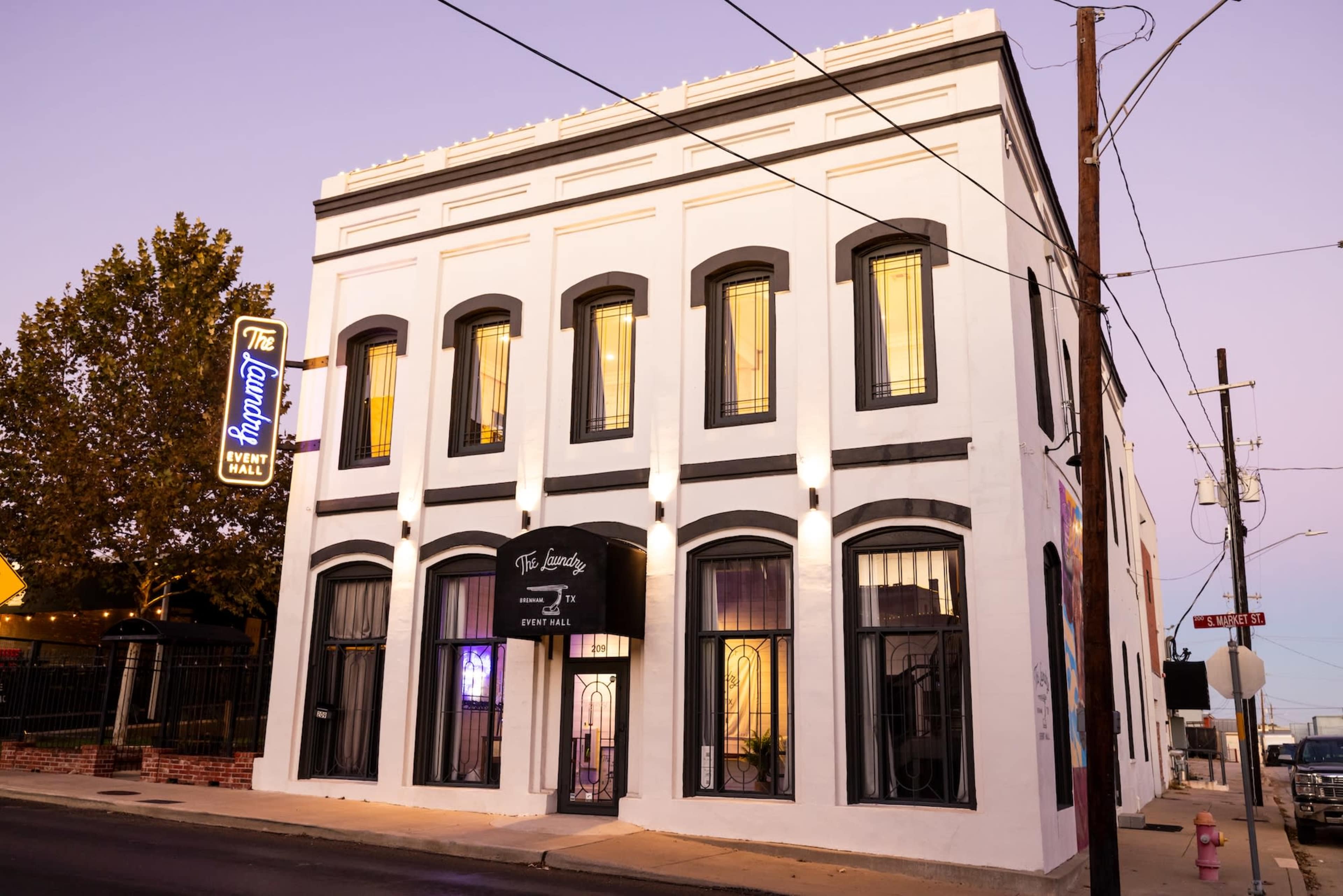 The image shows a white building with large windows and an illuminated sign reading "The Laundry Event Hall" on a street corner during twilight.