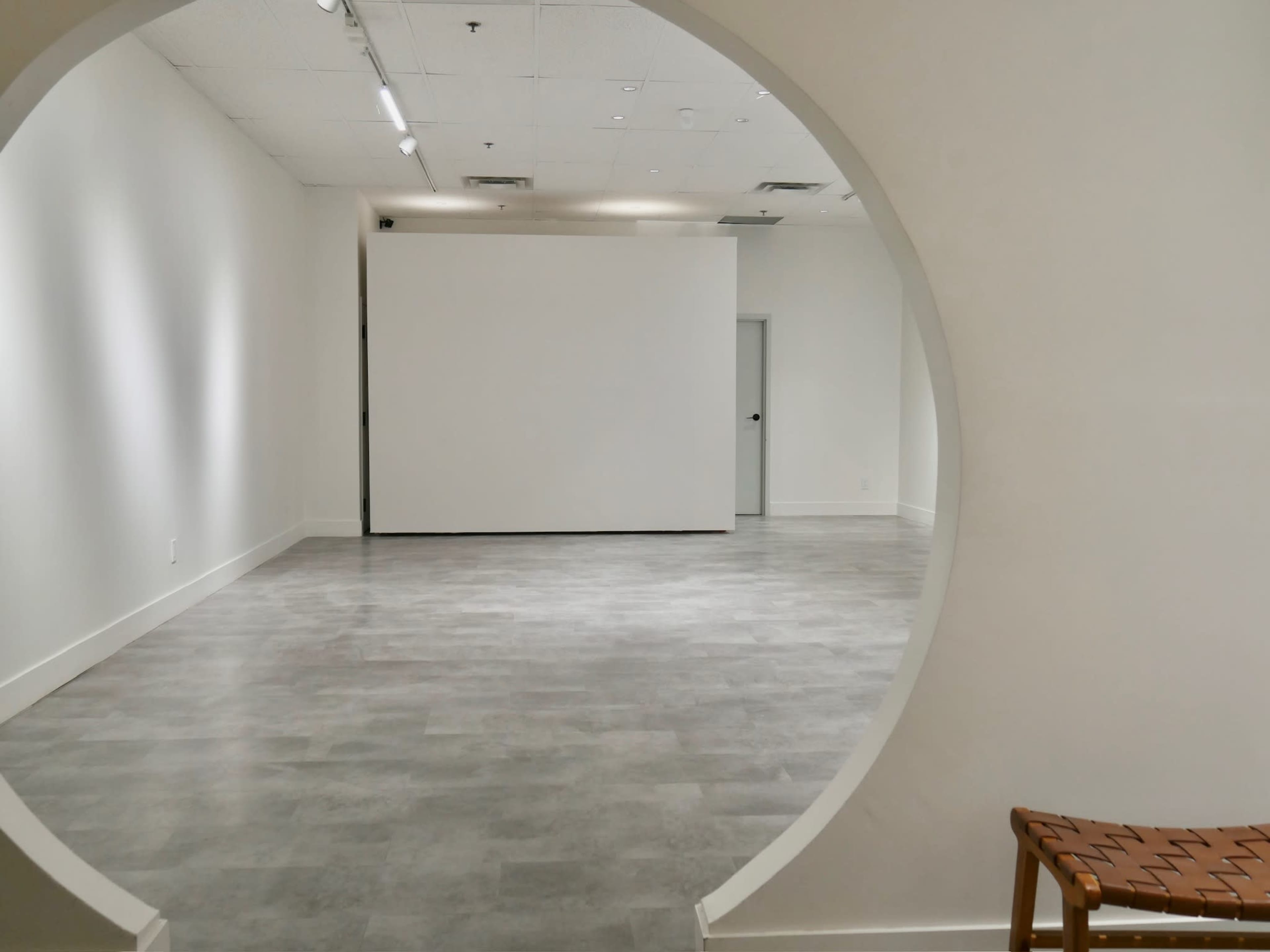 A minimalist interior space with gray flooring, a circular opening leading to a blank white wall, and a woven chair in the foreground.